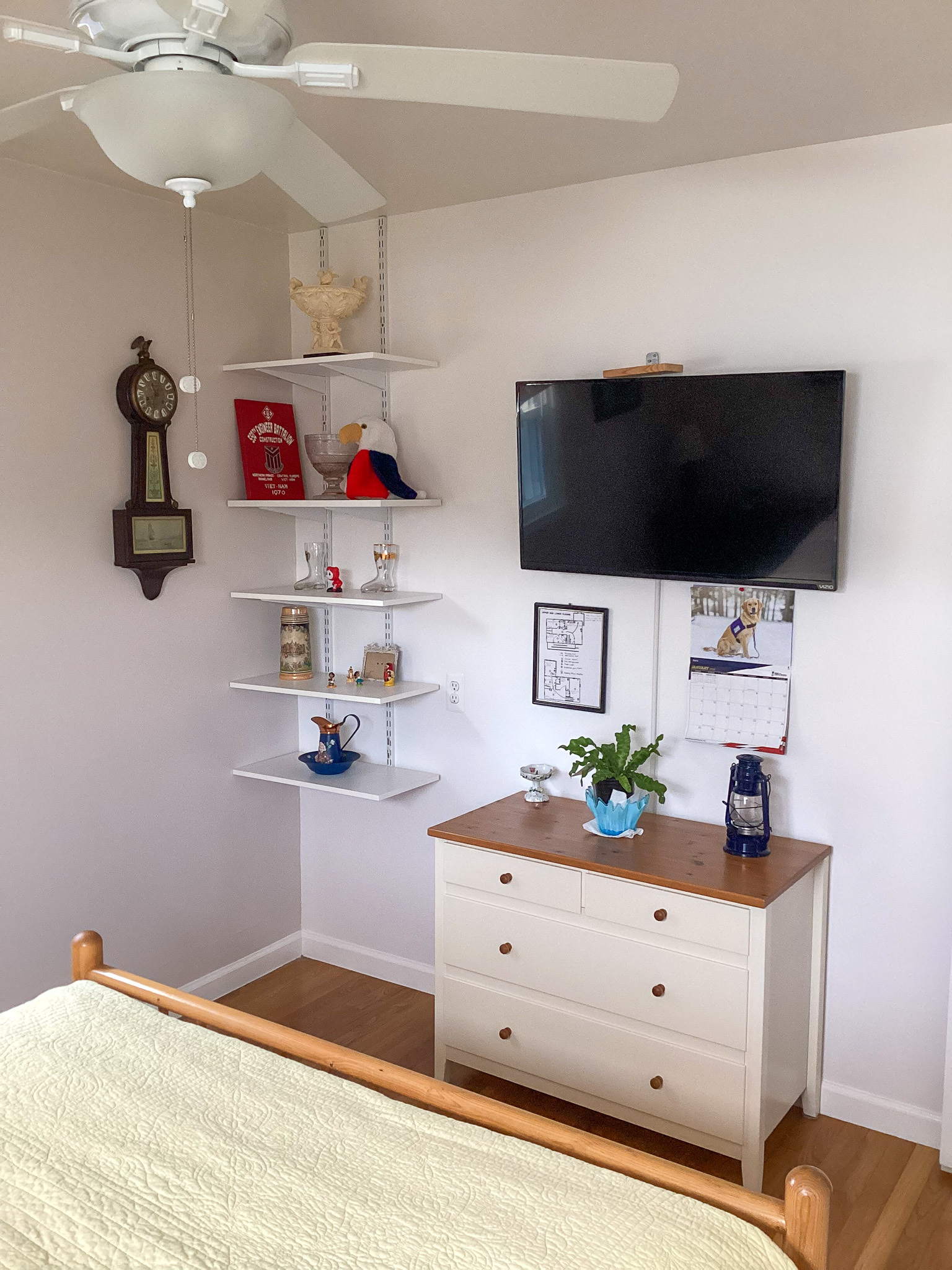 A bedroom corner with a white dresser topped with a plant and small items, a wall‑mounted TV above it, and open shelves displaying decorations beside a vintage-style clock, inside the home of Sponsored Residential Providers Teresa and Billy Herran in Woodbridge, Virginia.