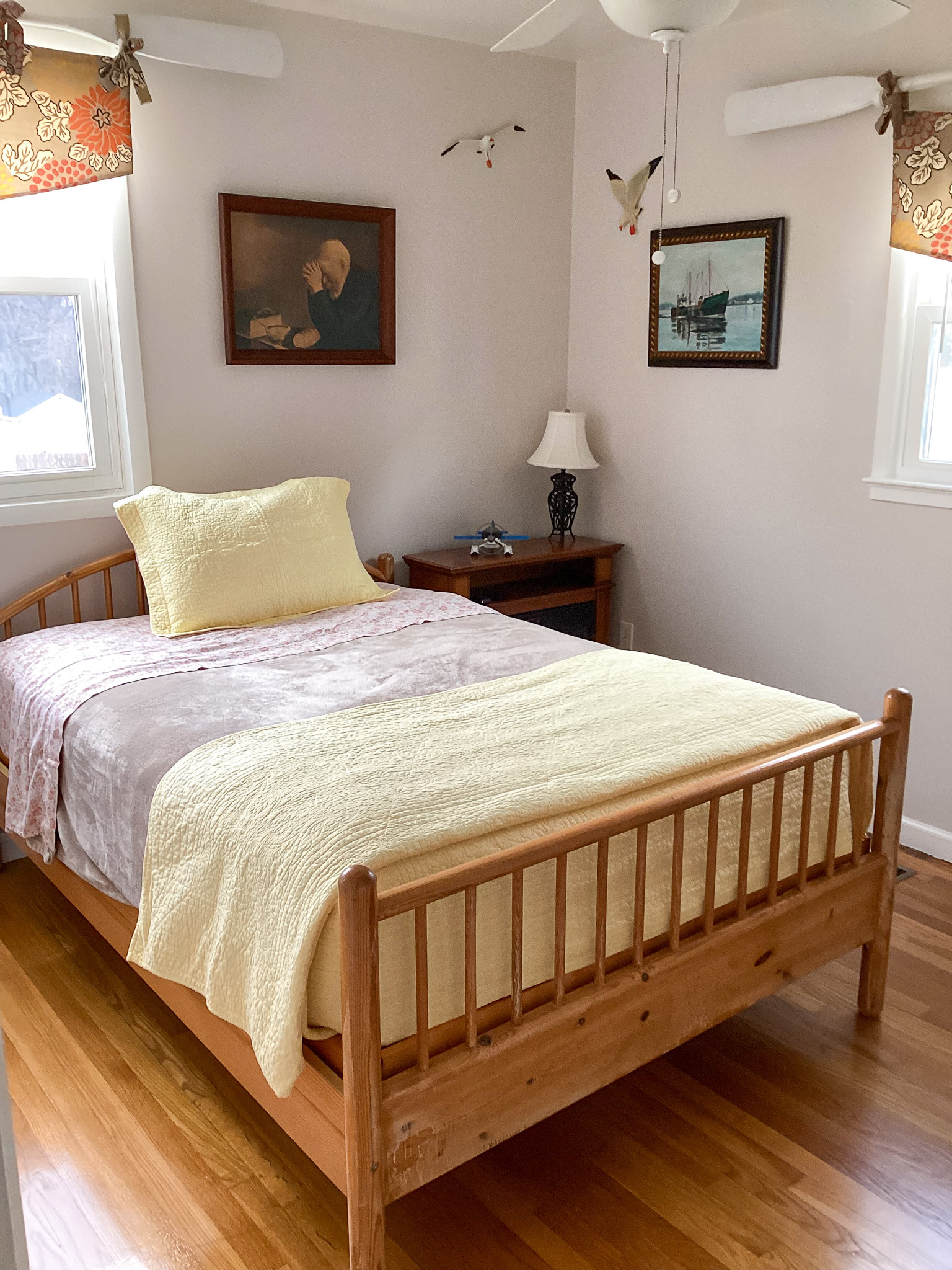A neatly made bedroom with a wooden bed frame, light yellow bedding, two windows with valances, framed artwork on the walls, and a bedside table with a lamp, inside the home of Sponsored Residential Providers Teresa and Billy Herran in Woodbridge, Virginia.