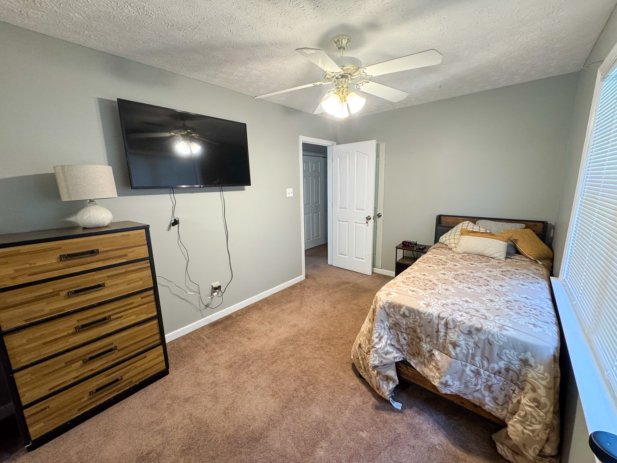 A cozy bedroom features a bed with patterned bedding, a dresser with a lamp, and a wall‑mounted TV above neatly arranged cords inside the home of Sponsored Residential Provider Durand Holloway in Chester, Virginia.