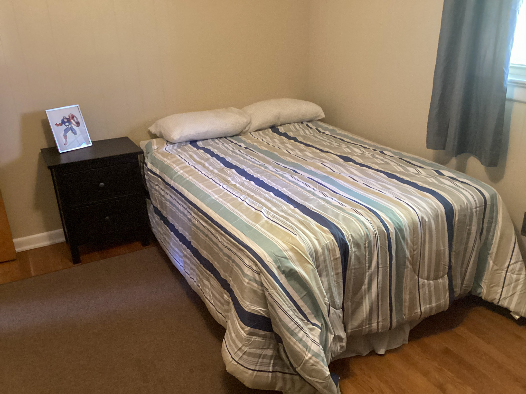 Simple bedroom with a bed covered in a striped comforter, two pillows, a dark nightstand with a framed photo, hardwood flooring, and a window with gray curtains inside the home of Sponsored Residential Providers Bill and Heather Terry in Staunton, Virginia.