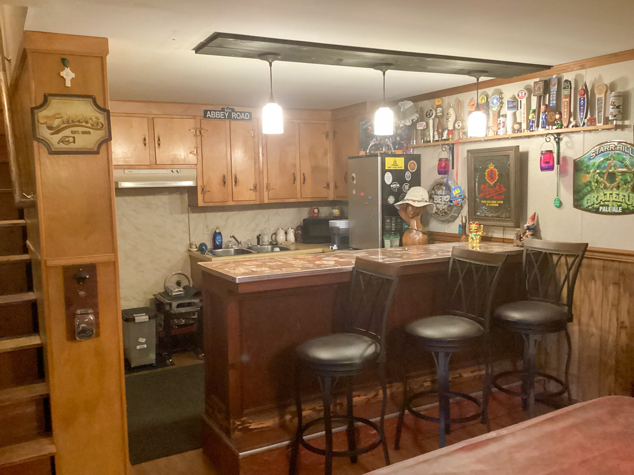 Basement bar area with wooden cabinetry, a countertop with three barstools, pendant lights overhead, a mini‑fridge covered with magnets, and assorted decor and signs on the walls inside the home of Sponsored Residential Providers Bill and Heather Terry in Staunton, Virginia.