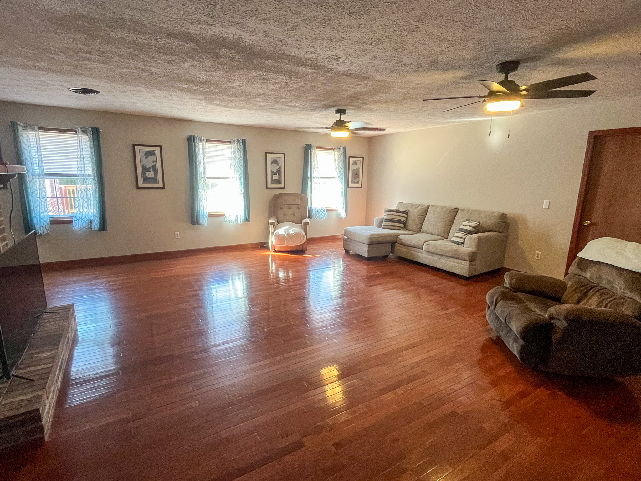 Bright living room with hardwood floors, three front-facing windows with blue curtains, ceiling fans, and light-colored seating arranged along the walls inside the home of Sponsored Residential Providers Penny Day and Steven Coleman in Hillsville, Virginia.