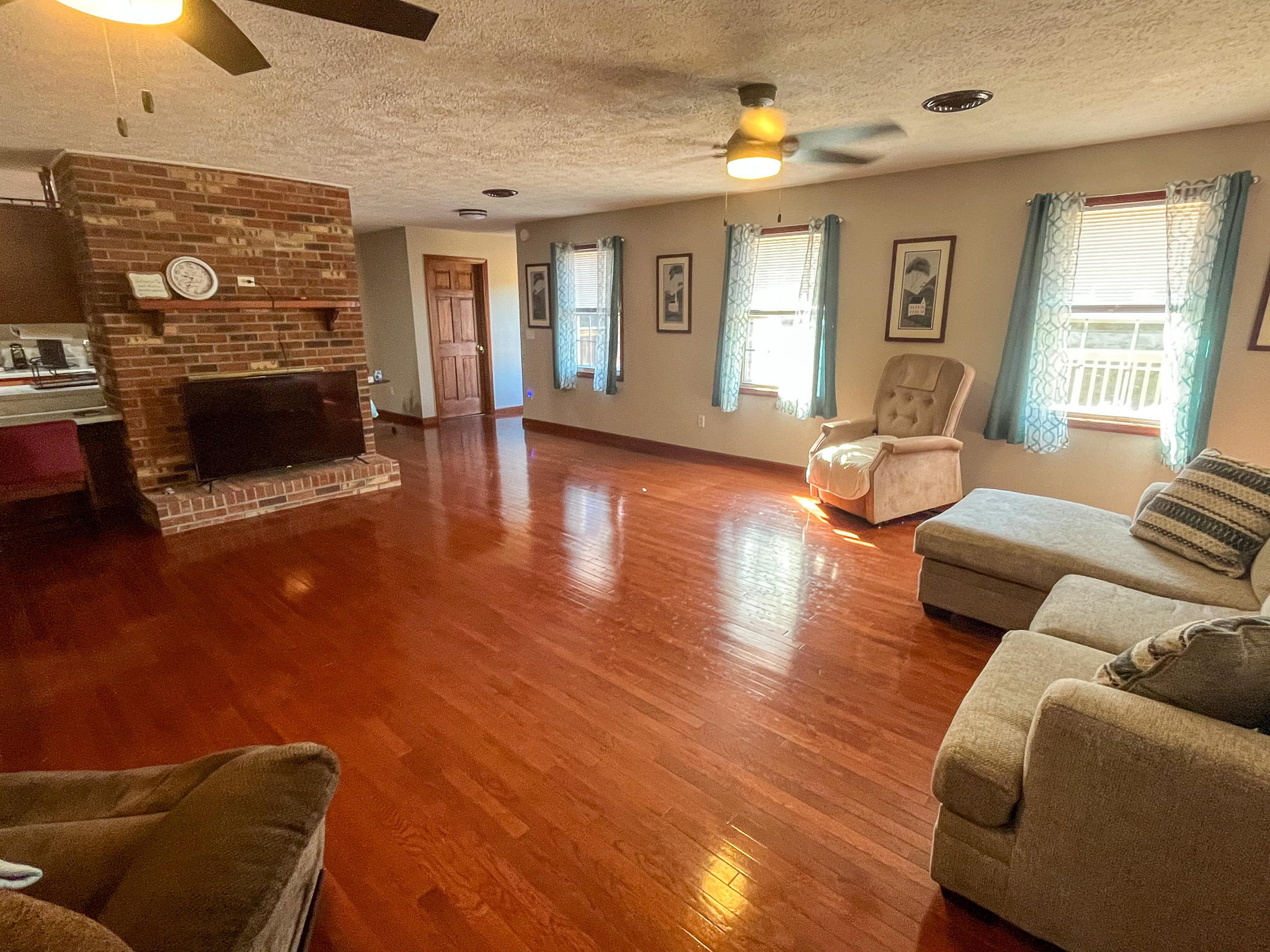 Spacious living room with polished hardwood floors, multiple windows with blue curtains, a brick fireplace, ceiling fans, and light-colored furniture inside the home of Sponsored Residential Providers Penny Day and Steven Coleman in Hillsville, Virginia.