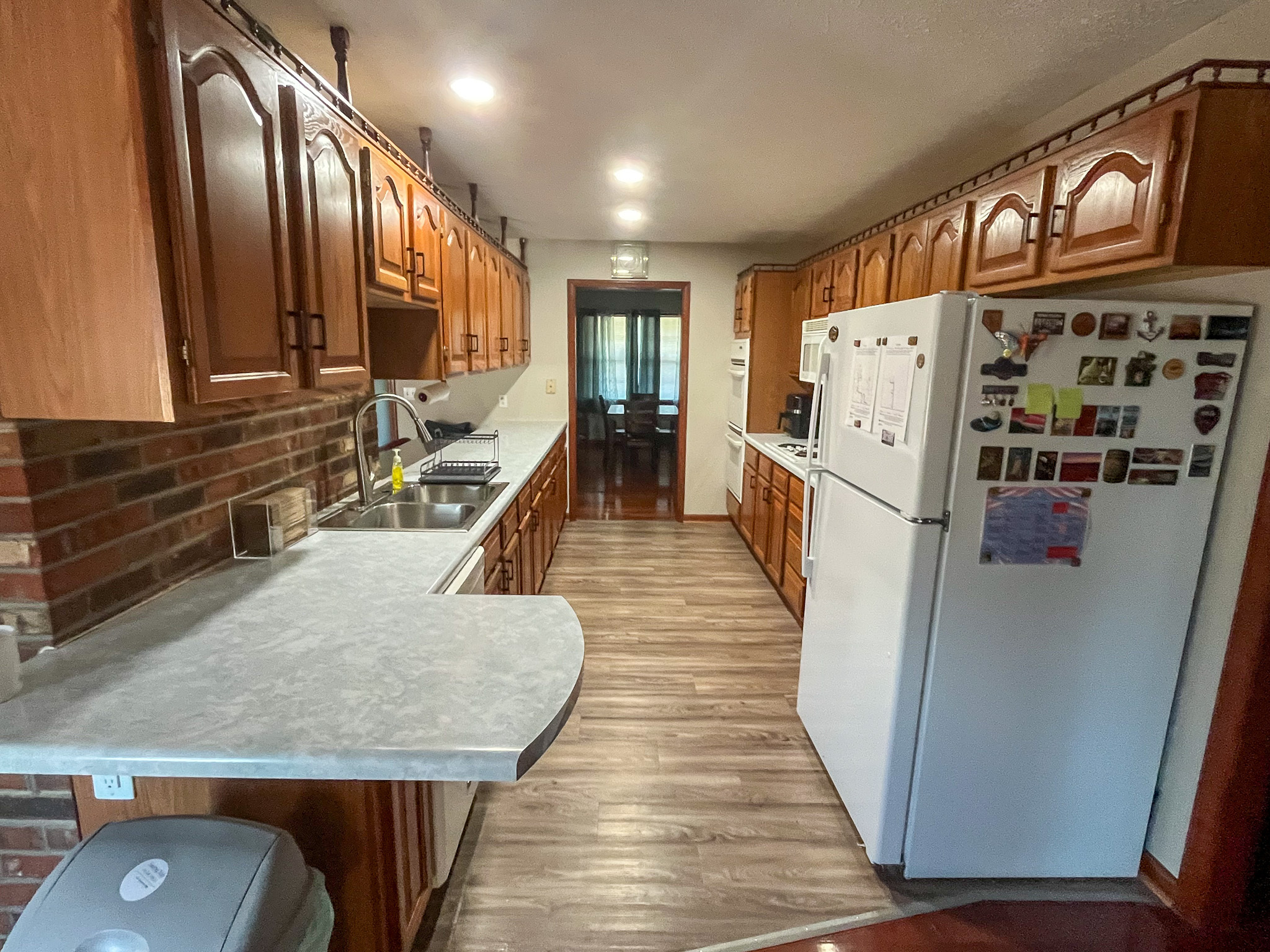 Galley-style kitchen with wood cabinets, brick backsplash, white appliances, long countertops, and wood-look flooring leading to a dining area inside the home of Sponsored Residential Providers Penny Day and Steven Coleman in Hillsville, Virginia.