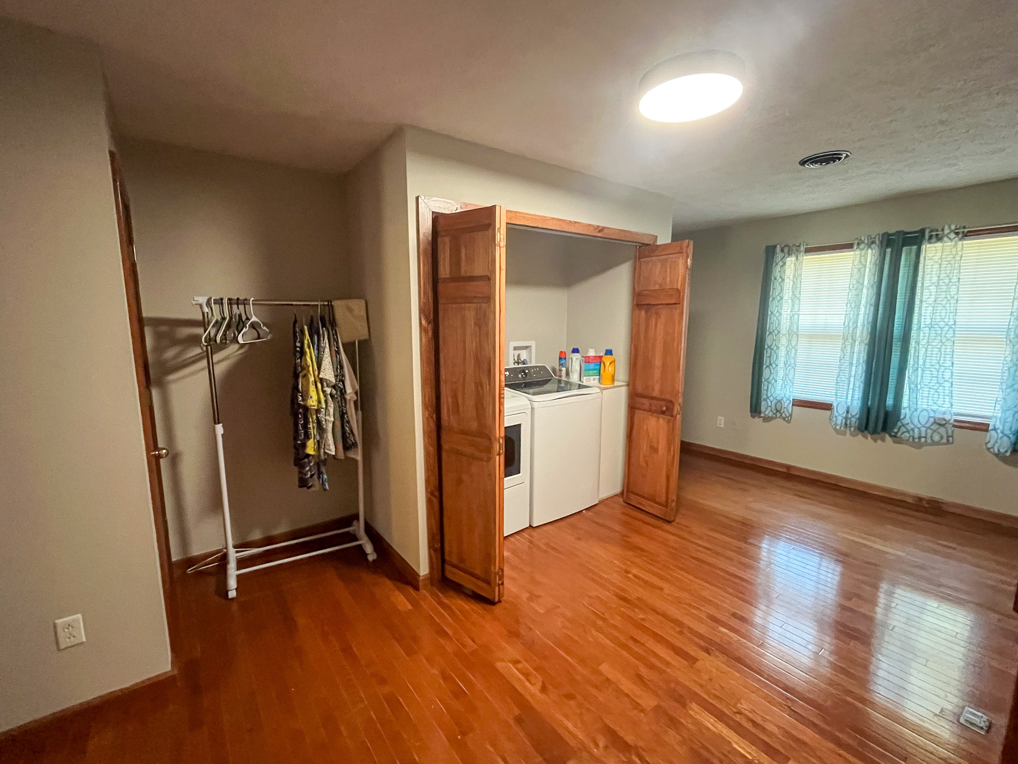 Room with hardwood floors, a window with blue curtains, an open closet containing a washer and dryer, and a clothing rack with hanging clothes inside the home of Sponsored Residential Providers Penny Day and Steven Coleman in Hillsville, Virginia.