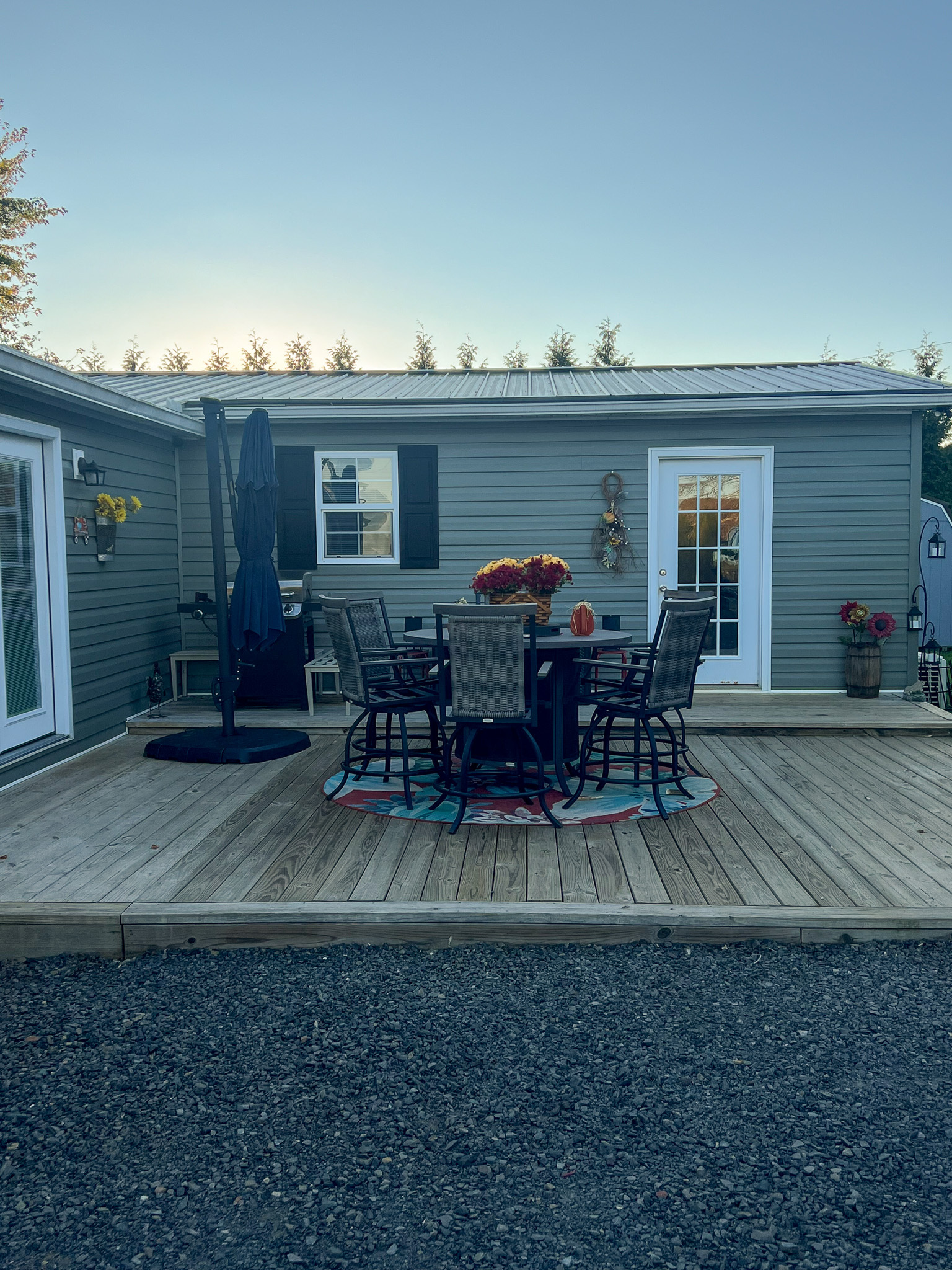 A wooden deck with a tall dining table and chairs, an umbrella, and potted plants, set against a gray house with windows and a glass door at dusk inside the home of Sponsored Residential Provider Christina Bedsaul in Hillsville, Virginia.