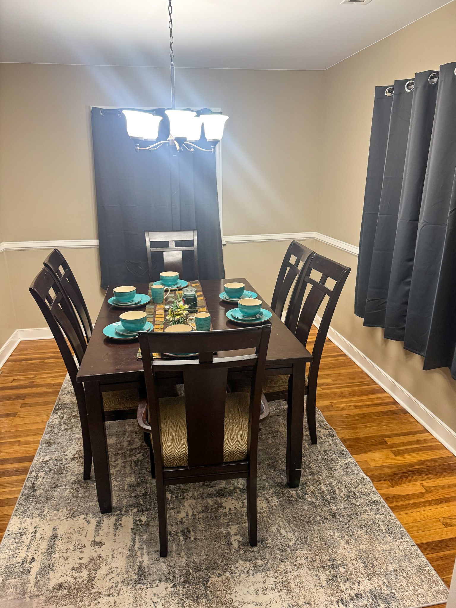 A dining room with a dark wood table set for six, teal place settings, a patterned rug, dark curtains, and a bright overhead light above the table inside the home of Sponsored Residential Provider Selena Banks in Chesterfield, Virginia.