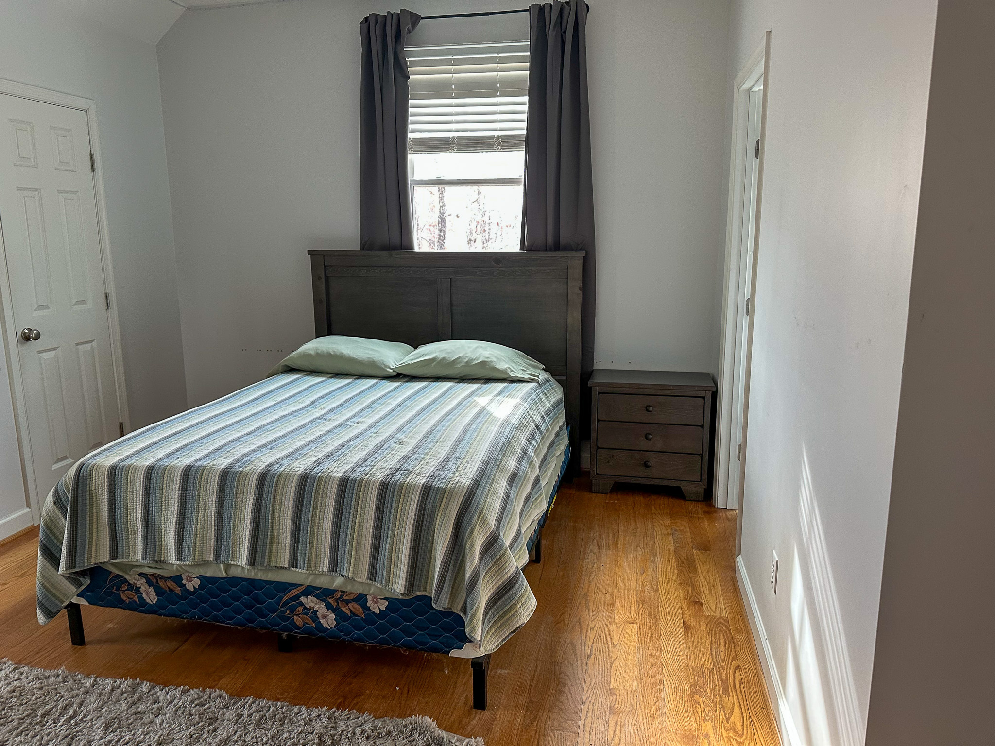 Simple bedroom with a neatly made bed, striped bedspread, dark headboard, bedside table, and a window with curtains above the bed inside the home of  Group Home Providers Felicia and Jason Martin in Amherst, Virginia.