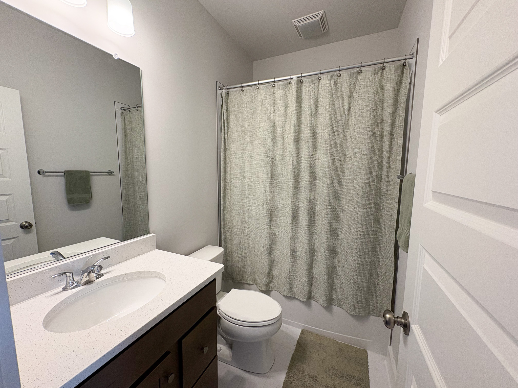 A clean bathroom with a white vanity, large mirror, toilet, and a shower with a light-colored patterned curtain, accented by matching towels and a floor mat inside the home of Sponsored Residential Providers Tania and LeSean Matthews in Glen Allen, Virginia.