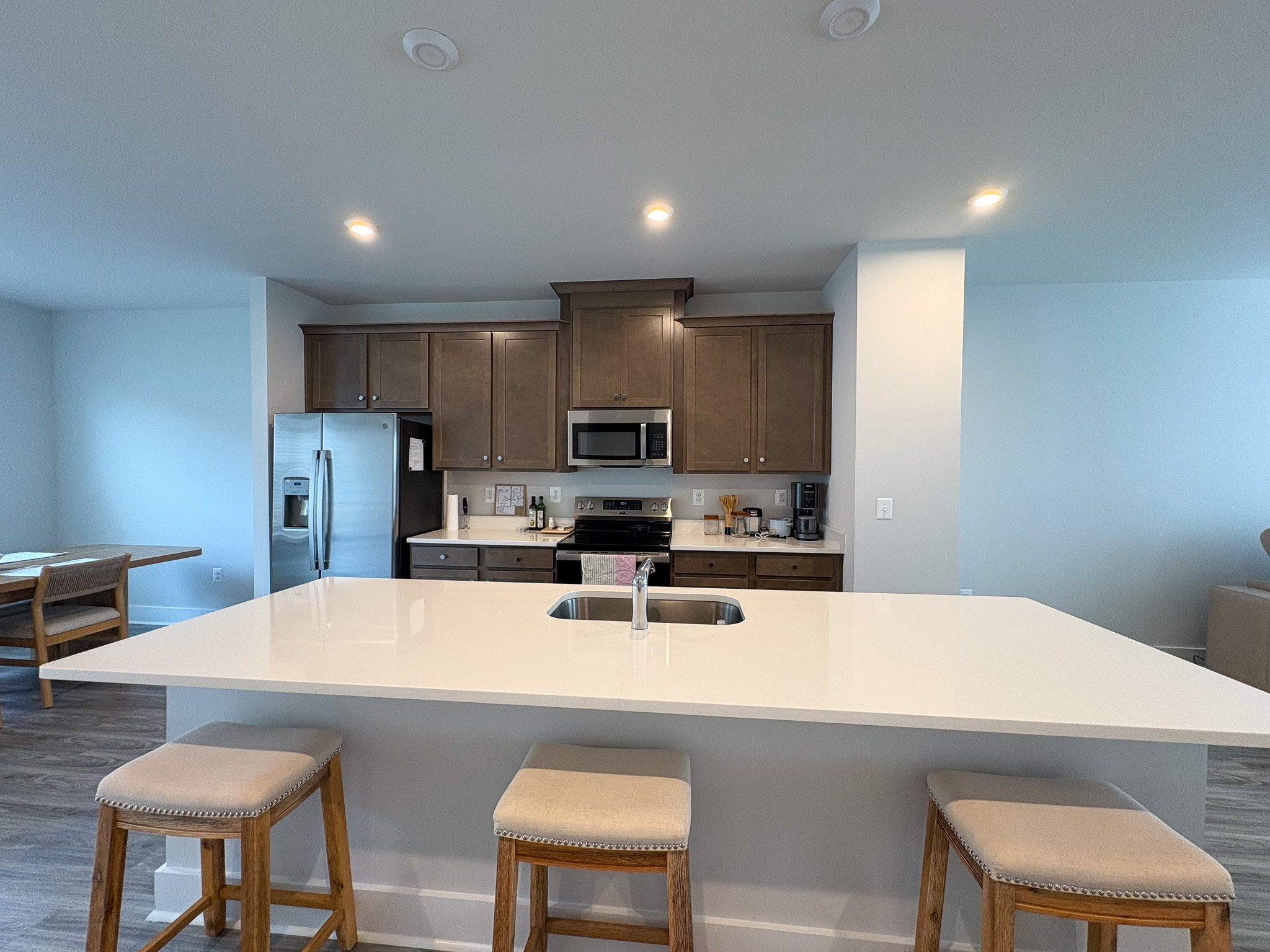 A modern kitchen with a large white island featuring three wooden stools, stainless steel appliances, wood‑tone cabinets, and recessed ceiling lights inside the home of Sponsored Residential Providers Tania and LeSean Matthews in Glen Allen, Virginia.