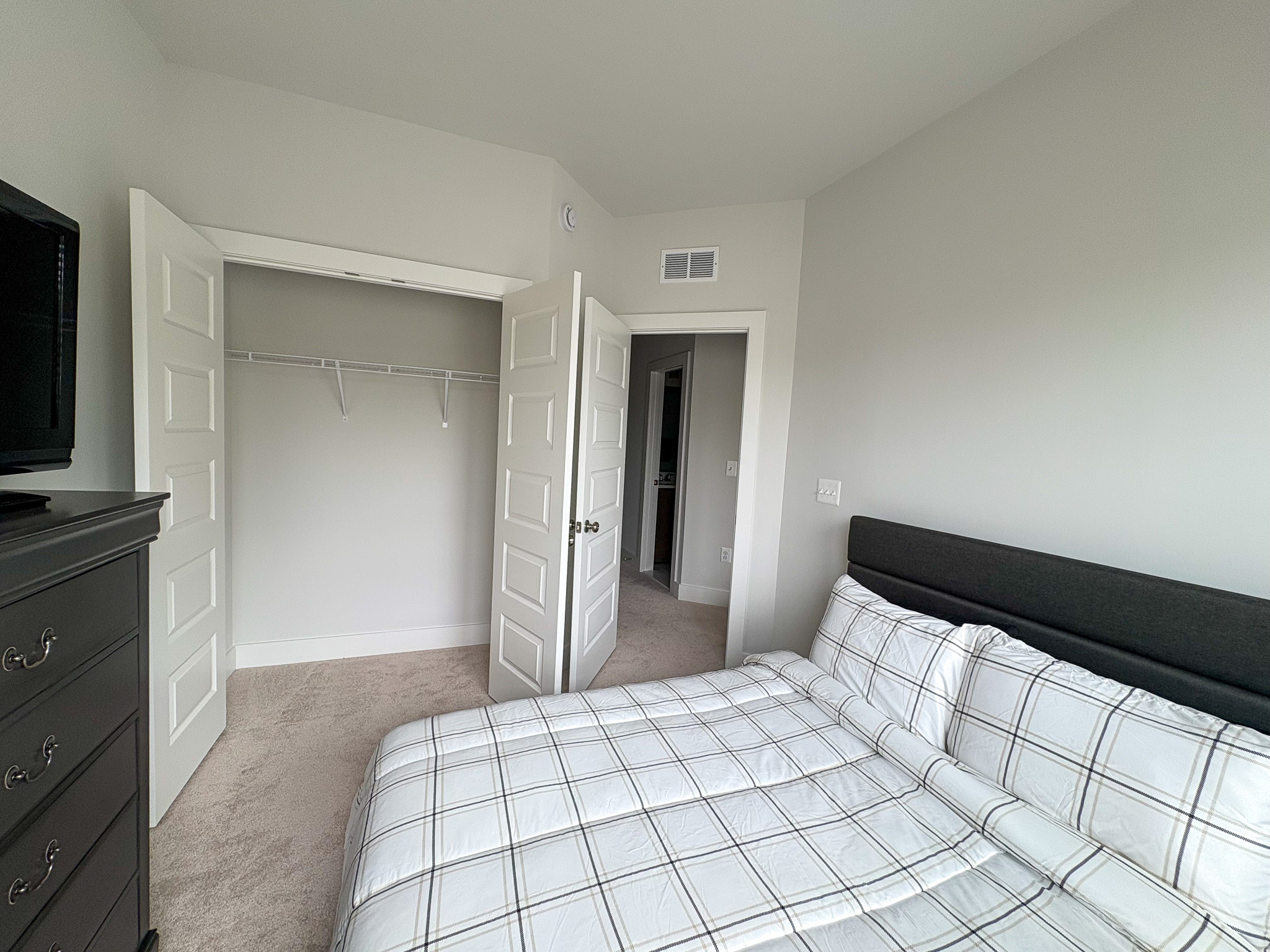 A bright bedroom with a plaid‑patterned bed, a dark dresser with a TV on top, white double closet doors, and a doorway leading to the hall inside the home of Sponsored Residential Providers Tania and LeSean Matthews in Glen Allen, Virginia.