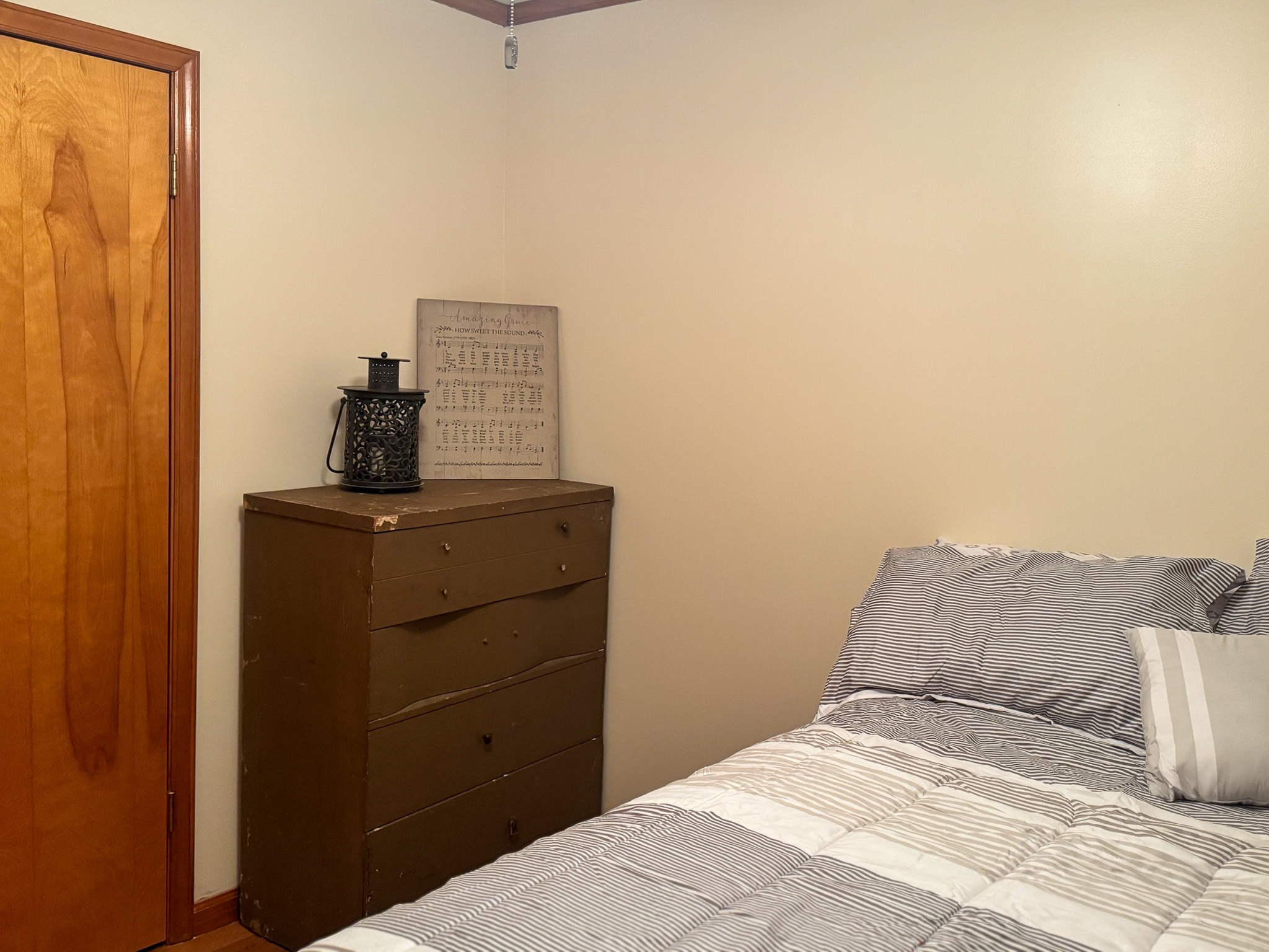 A bedroom corner with a brown dresser topped with decor and a framed sign, a wooden door on the left, and part of a bed with gray and white bedding on the right inside the home of Sponsored Residential Provider Pamela Hill in Austinville, Virginia.