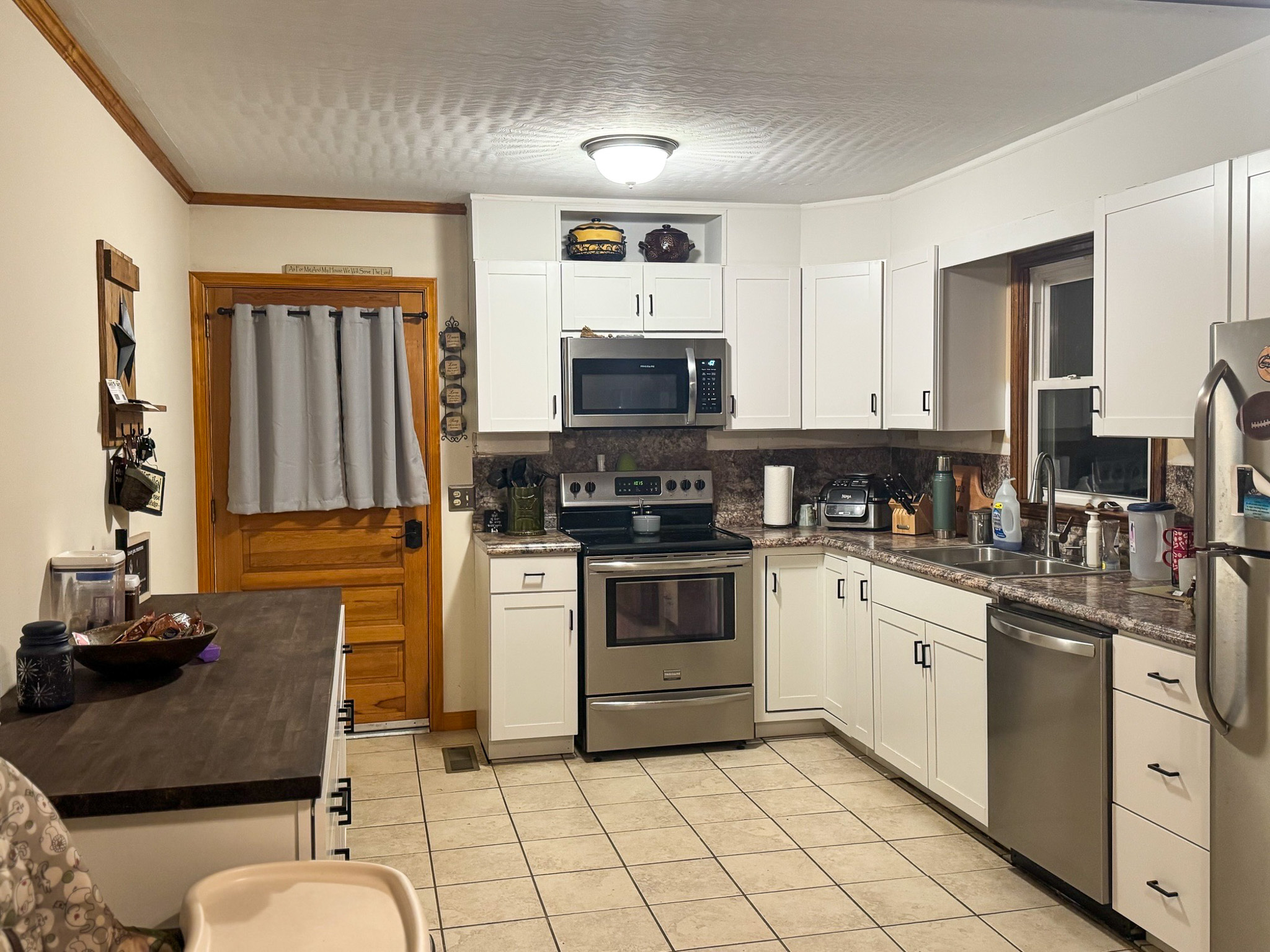 A kitchen with white cabinets, stainless steel appliances, tiled flooring, a wooden door with a curtain, and a long counter on the left with shelves above it inside the home of Sponsored Residential Provider Pamela Hill in Austinville, Virginia.