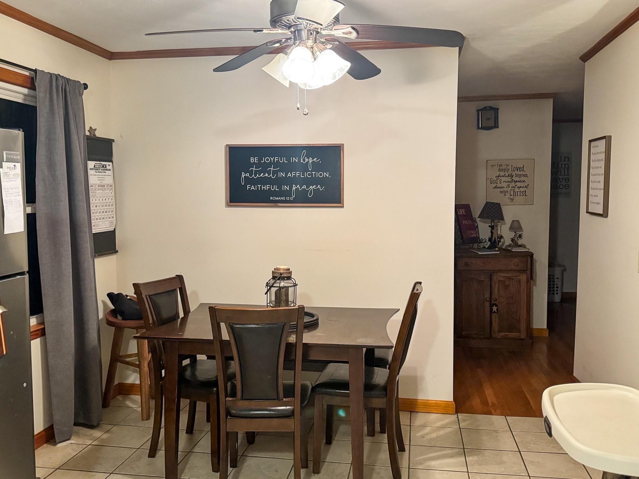 A dining area with a dark wood table and four chairs, a ceiling fan with lights above, a chalkboard on the wall, tile flooring, and a small hallway leading to additional rooms inside the home of Sponsored Residential Provider Pamela Hill in Austinville, Virginia.
