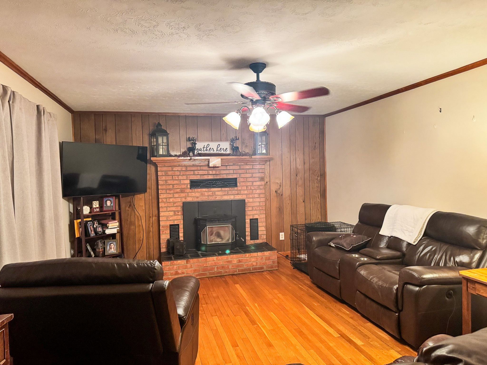 A living room with wood‑paneled walls, a brick fireplace, dark leather seating, a ceiling fan with lights, a wall‑mounted TV on the left, and hardwood flooring inside the home of Sponsored Residential Provider Pamela Hill in Austinville, Virginia.