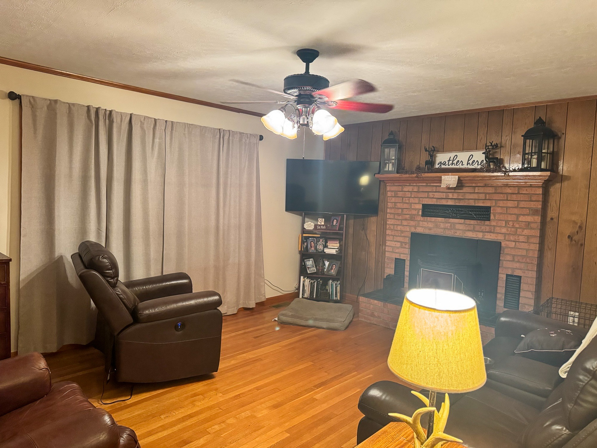 A living room with wood‑paneled walls, a brick fireplace, dark leather recliners, a ceiling fan with lights, a floor lamp with a yellow shade, and hardwood floors inside the home of Sponsored Residential Provider Pamela Hill in Austinville, Virginia.