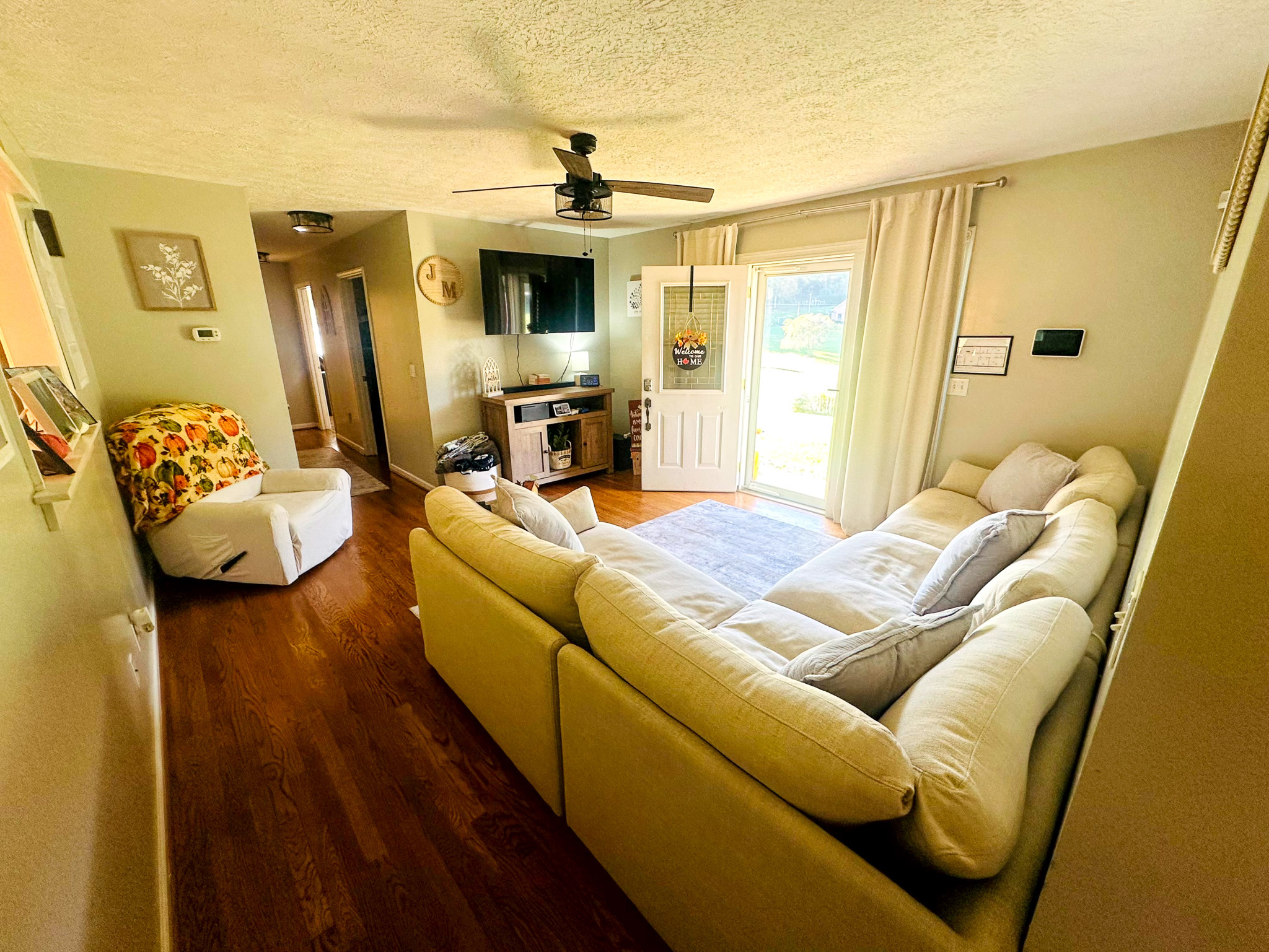 A bright living room with light green walls, a large cream sectional sofa, wood floors, a wall‑mounted TV above a small console, and sunlight streaming through a glass front door with curtains inside the home of Sponsored Residential Provider Jayda Utt in Galax, Virginia.