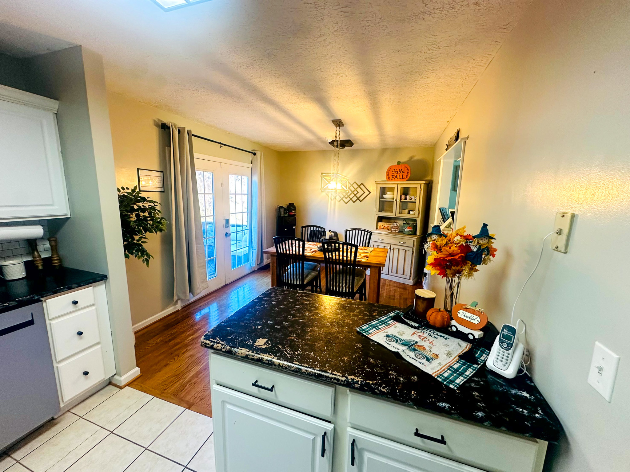 A view from the kitchen over black granite counters toward a dining area with wood floors, French doors, a hanging light fixture, and a small shelving unit decorated with seasonal items inside the home of Sponsored Residential Provider Jayda Utt in Galax, Virginia.