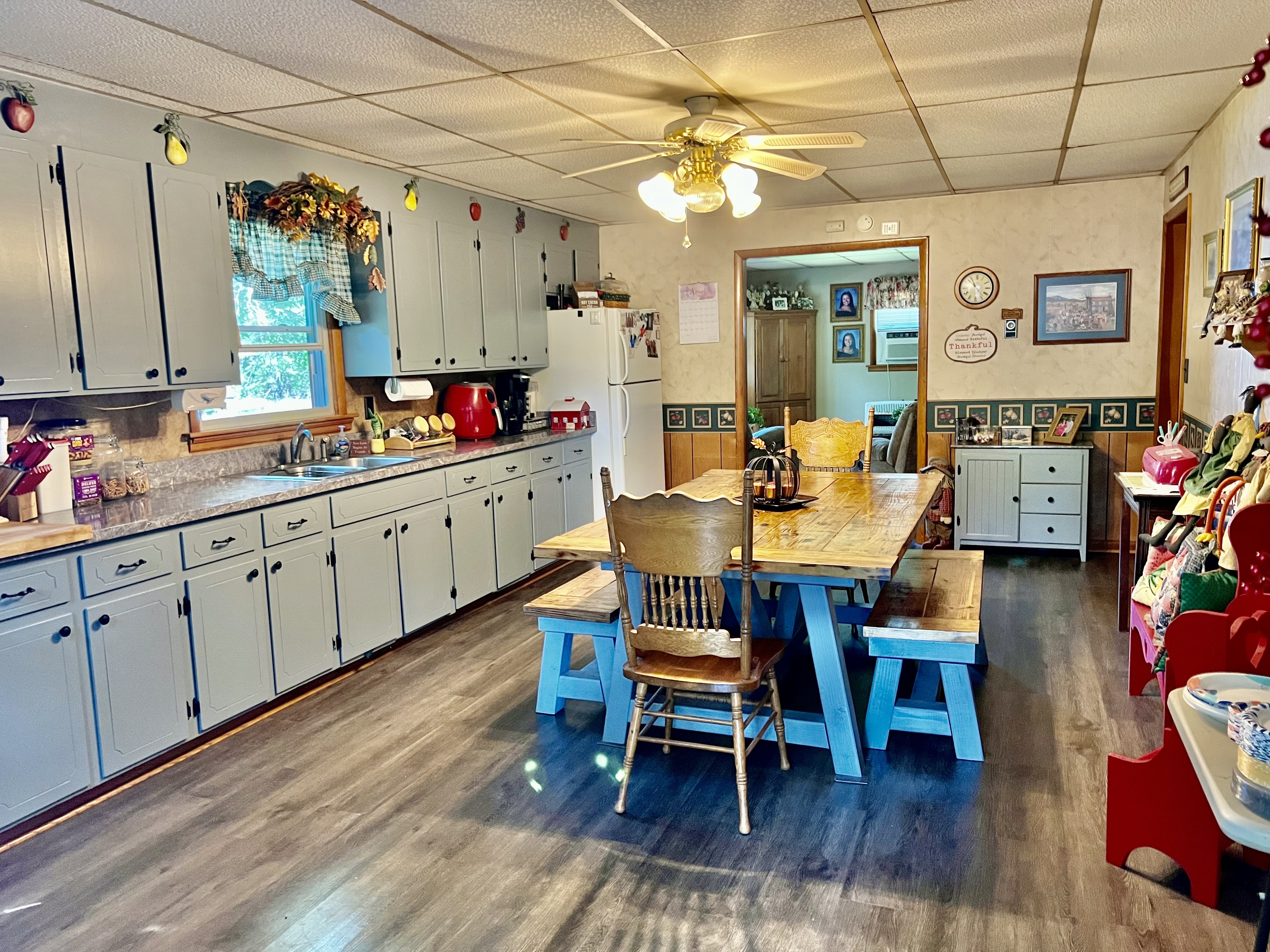A spacious kitchen with light blue cabinets, a long wooden dining table with mixed chairs and benches, a ceiling fan, and decorative items along the counters and walls inside the home of Sponsored Residential Providers Beth and Otis Fowler in Rustburg, Virginia.