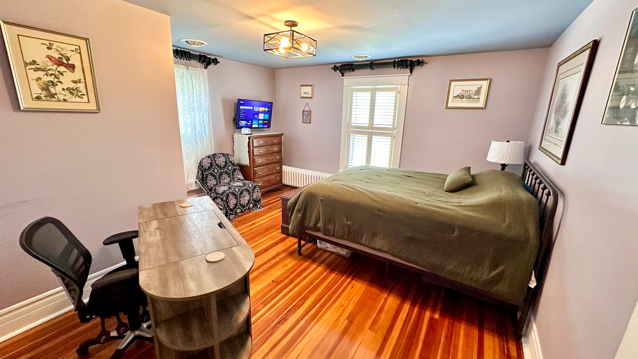 Bedroom with a desk and office chair, a bed with a green comforter, patterned armchair, wall art, and hardwood floors inside the home of Sponsored Residential Providers Jeff and Sarah McLane in Lynchburg, Virginia.