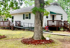 A white house with green shutters has two wooden porches connected by a long metal accessibility ramp, with a large tree in the foreground surrounded by a mulched bed belonging to Sponsored Residential Providers Beth and Otis Fowler in Rustburg, Virginia.