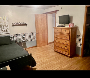 A bedroom with wood flooring, a twin bed with a black comforter, a tall wooden dresser with a TV on top, and a checkered accent border along the walls inside the home of Sponsored Residential Providers Beth and Otis Fowler in Rustburg, Virginia.
