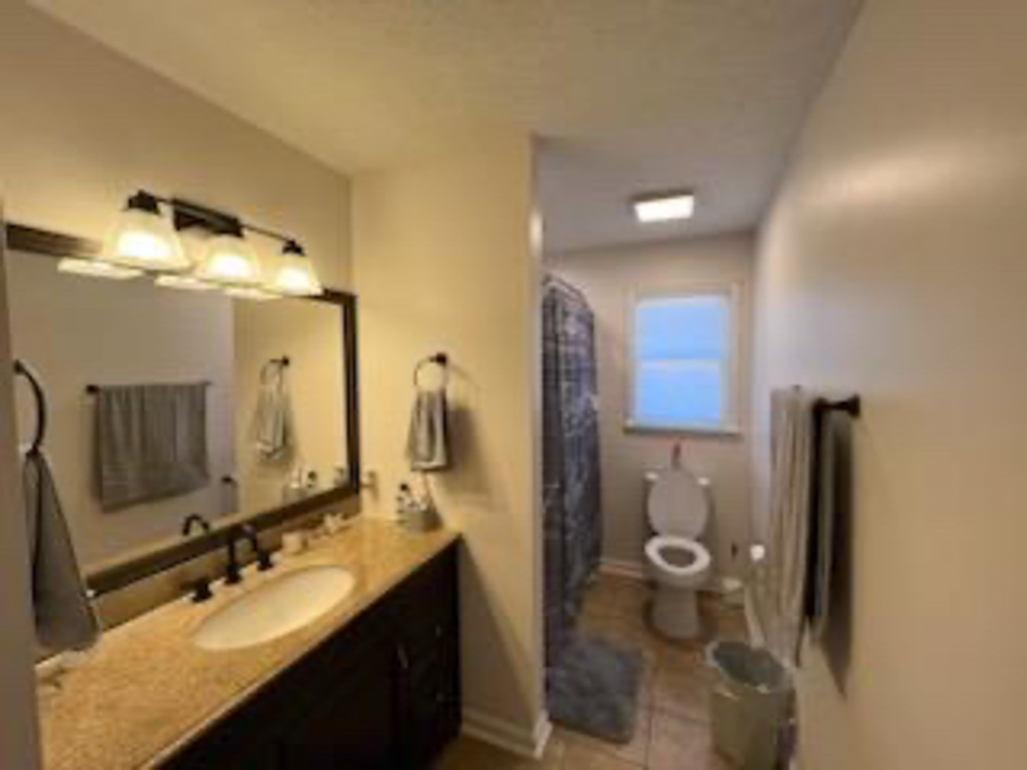 A bathroom with a long vanity and a lighted mirror on the left, leading to a toilet and a shower with a patterned curtain at the back, with beige walls and tile flooring inside the home of Sponsored Residential Provider Devonne Dixon in Lynchburg, Virginia.