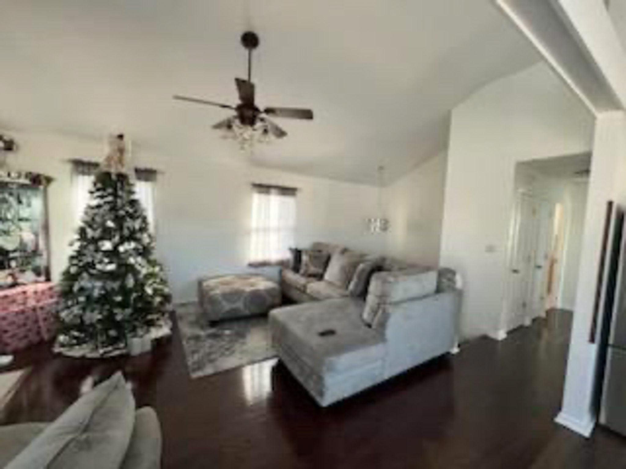 A living room with dark wood floors, light gray sectional sofas, a ceiling fan, and a decorated Christmas tree near the window inside the home of Sponsored Residential Provider Devonne Dixon in Lynchburg, Virginia.