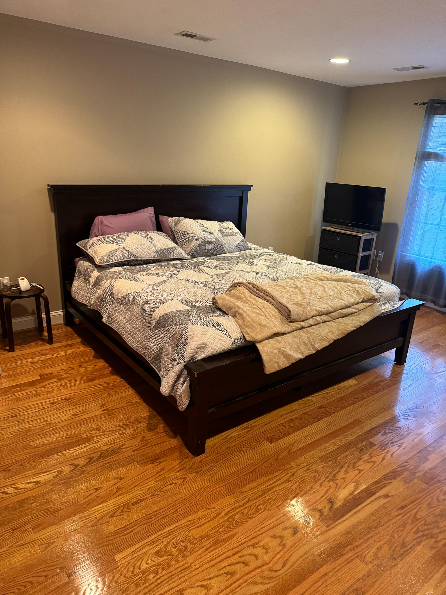 A bedroom with a dark wooden bed frame, light bedding, hardwood floors, and a TV stand near the window inside the home of Sponsored Residential Provider Sharon Jennings in Salem, Virginia.