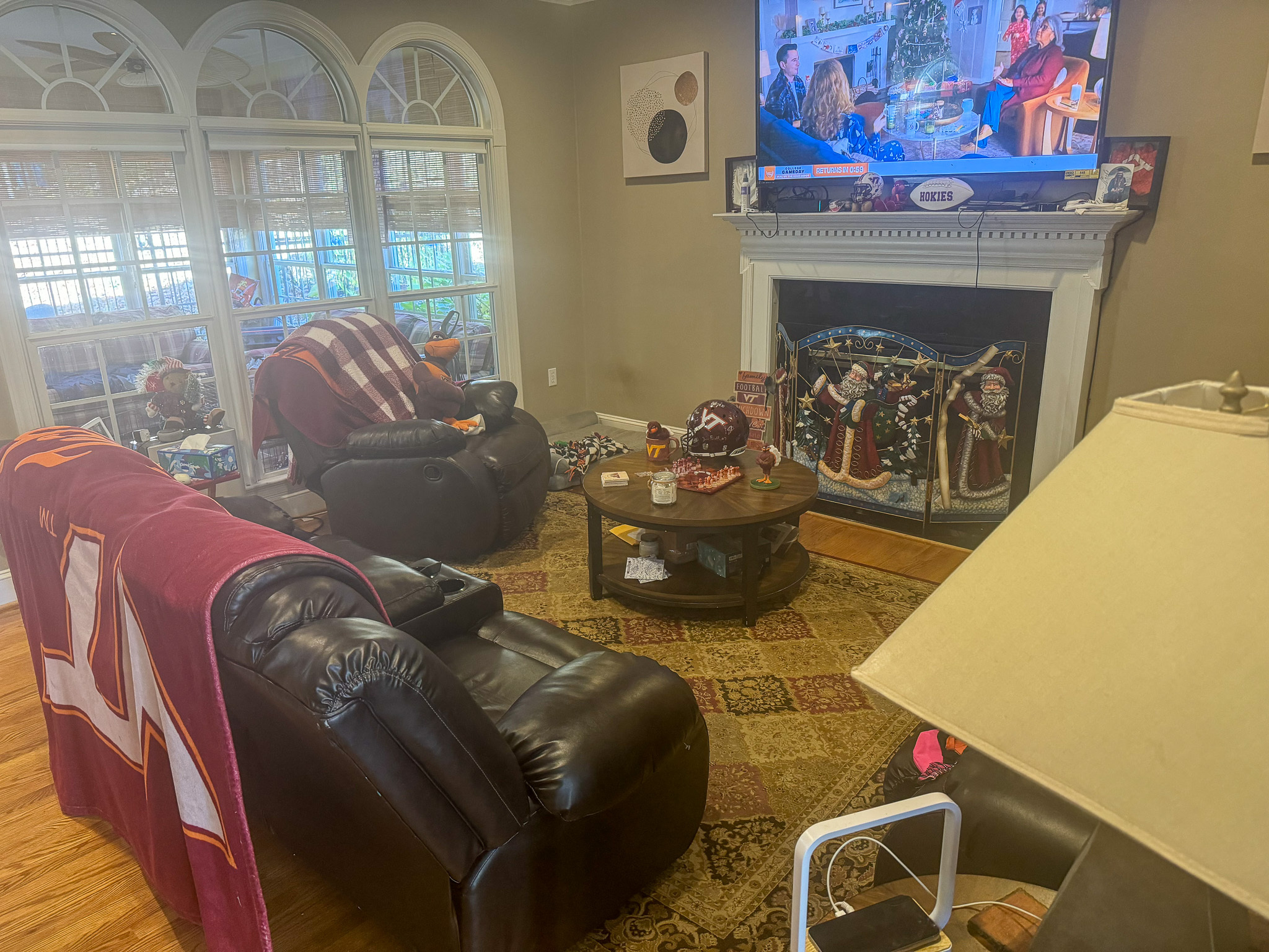 A living room with dark leather seating, a TV above a decorated fireplace, large arched windows, and a round coffee table inside the home of Sponsored Residential Provider Sharon Jennings in Salem, Virginia.
