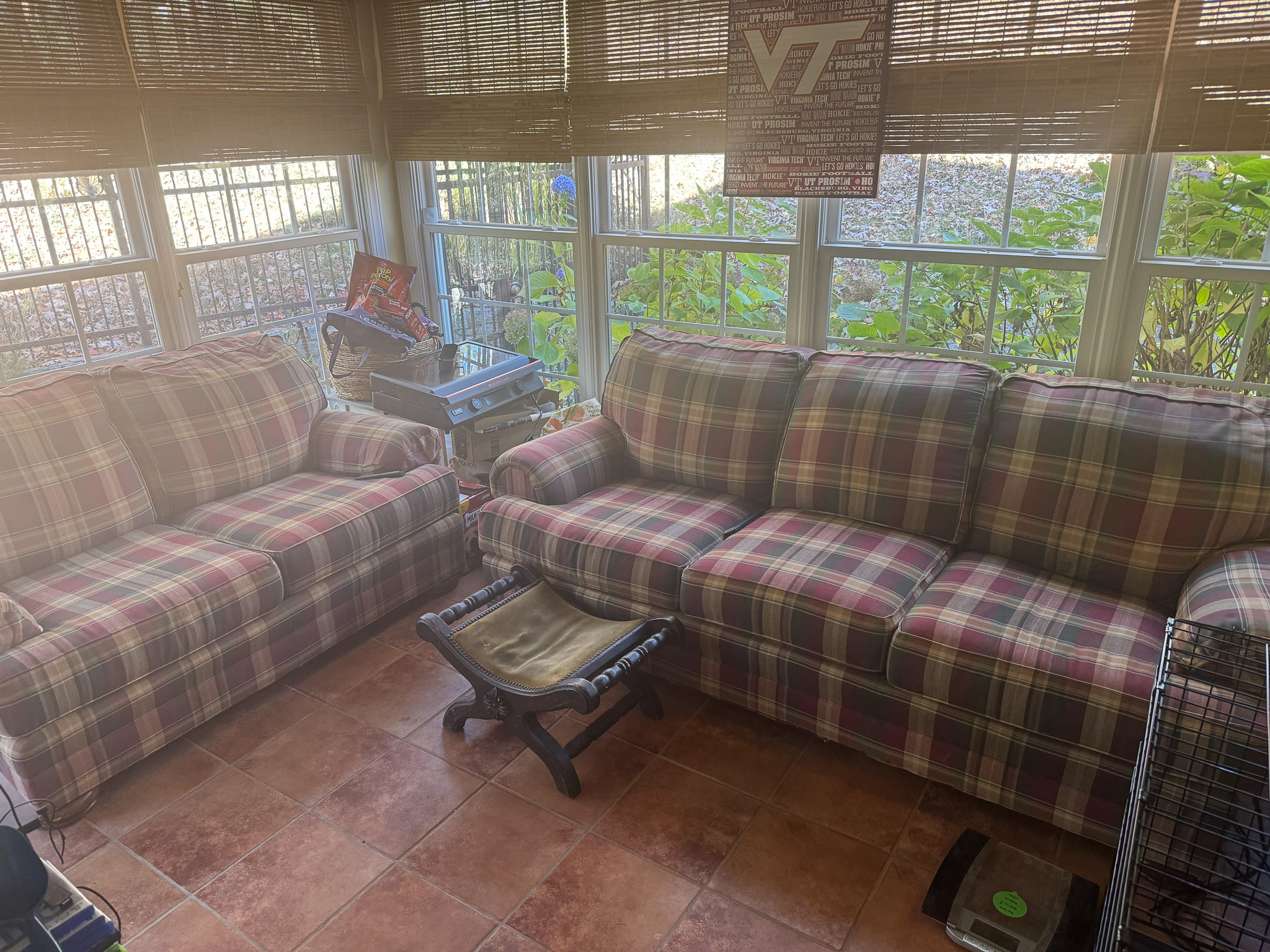 A sunroom with two plaid sofas, woven blinds, tiled flooring, and greenery visible through the surrounding windows inside the home of Sponsored Residential Provider Sharon Jennings in Salem, Virginia.