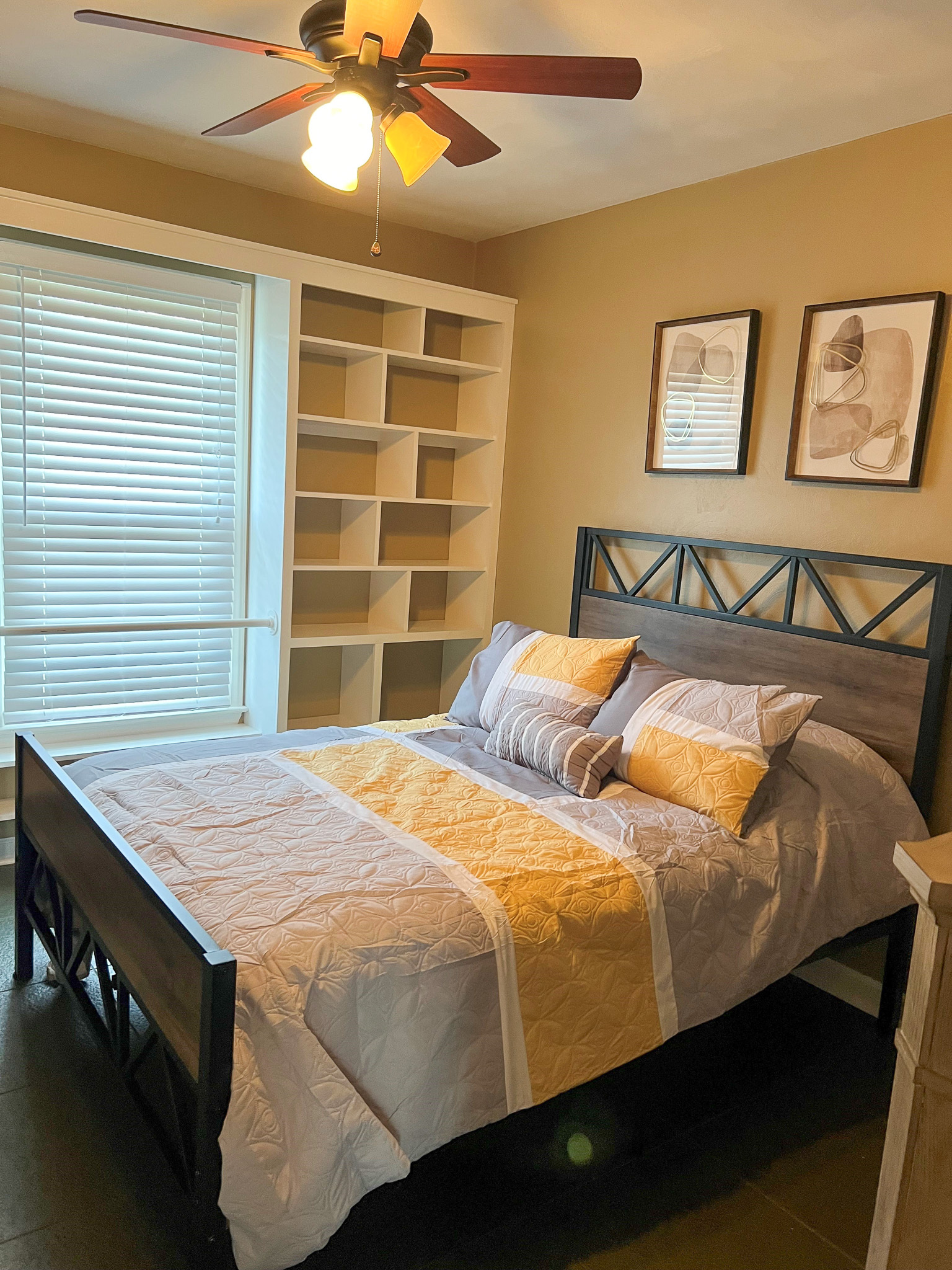 A bedroom with beige walls, a metal bed frame with yellow-accented bedding, built‑in shelves, and a ceiling fan with a light inside the home of Group Home Providers Anthony and Melissa Johnson in Daleville, Virginia.