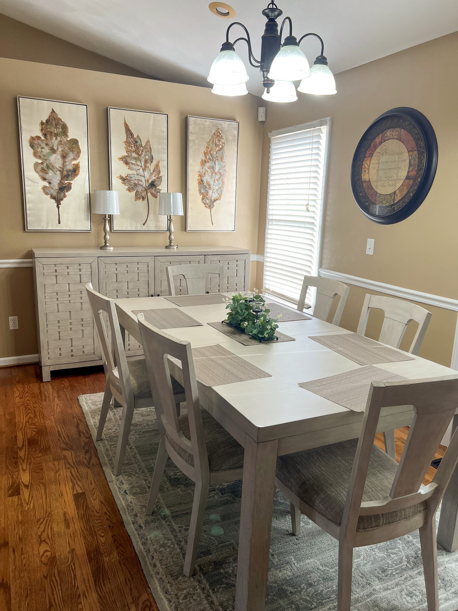 A dining room with a light wood table and chairs, a sideboard with lamps, botanical artwork, and a circular wall decor piece inside the home of Group Home Providers Anthony and Melissa Johnson in Daleville, Virginia.