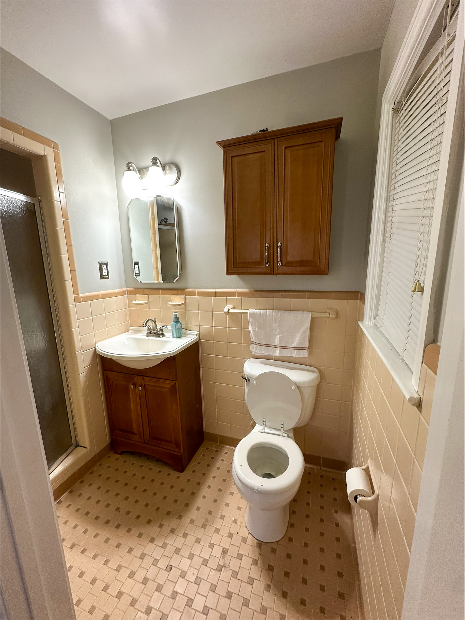 A small bathroom with a shower, a pedestal sink, a toilet, and wooden storage cabinets inside the home of Group Home Provider Tina Langhorn in Roanoke, Virginia.