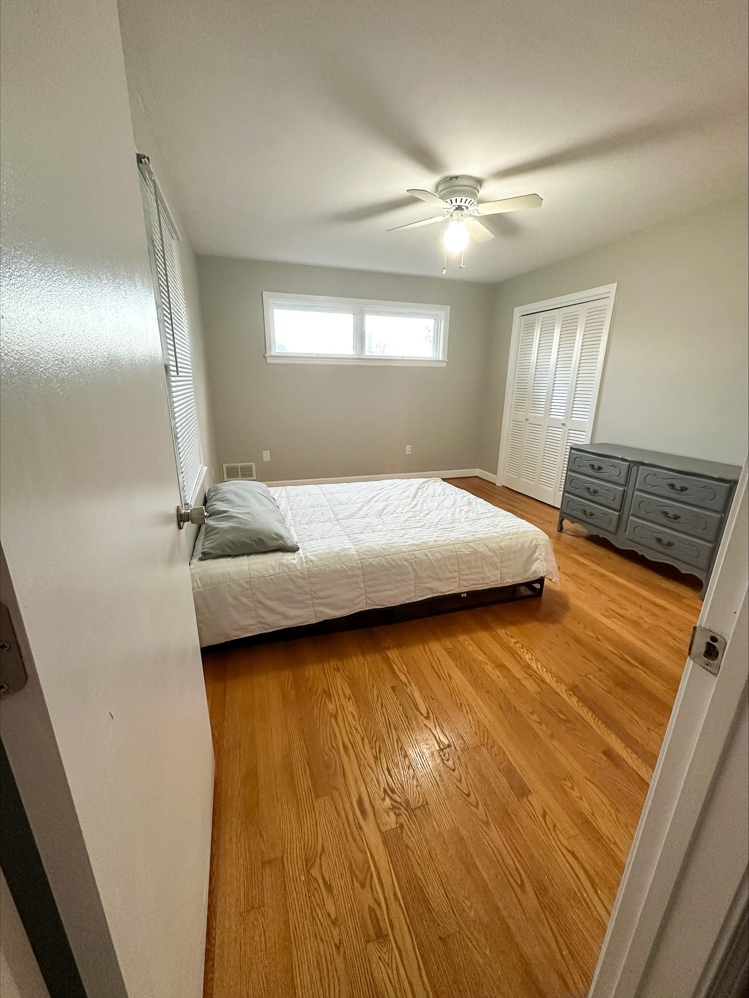 A simple bedroom with a low bed, hardwood floors, a ceiling fan, and a gray dresser beneath two small windows inside the home of Group Home Provider Tina Langhorn in Roanoke, Virginia.