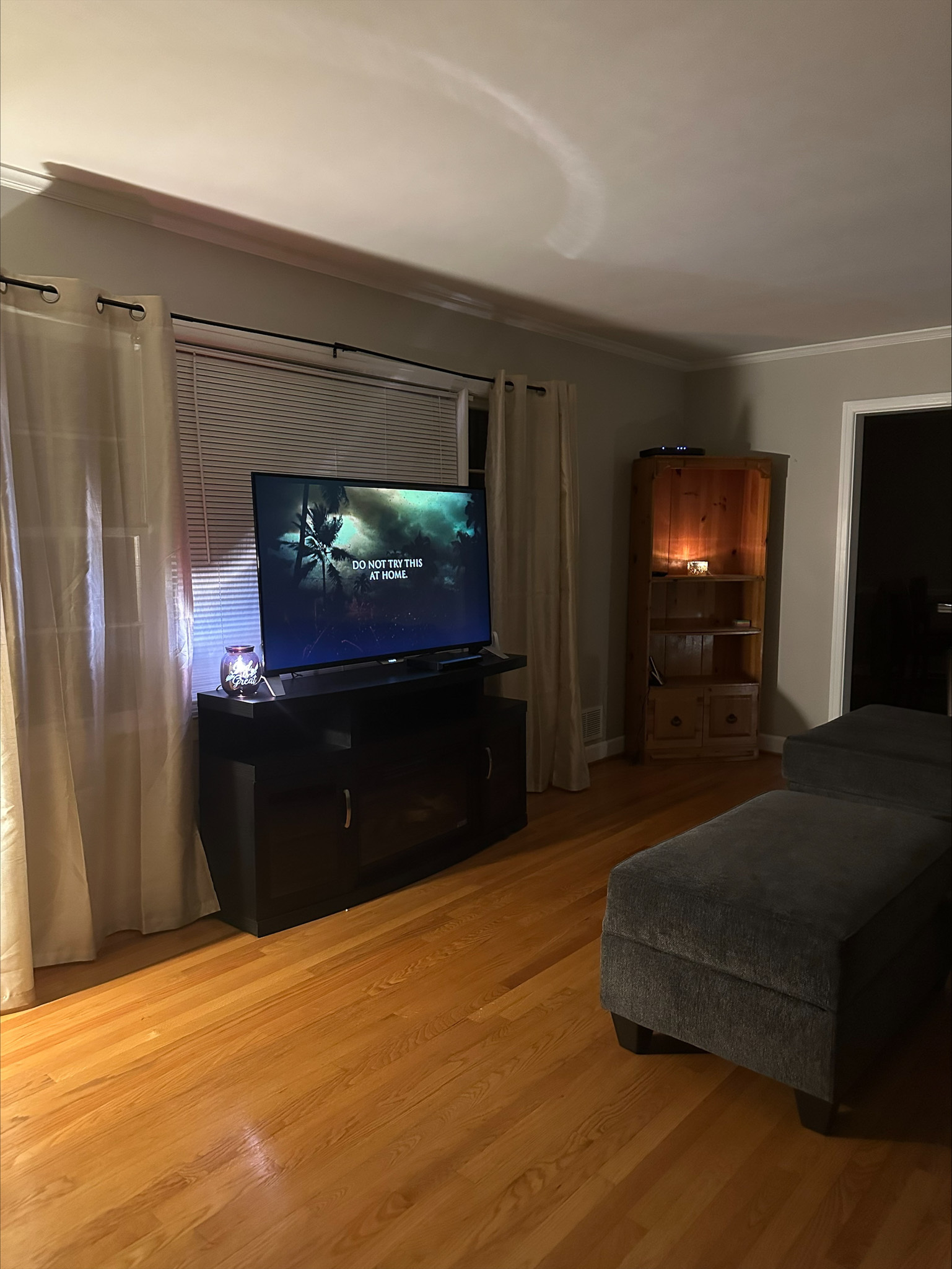 A dimly lit living room with a TV on a stand, long curtains behind it, a gray ottoman, and a lit corner cabinet inside the home of Group Home Provider Tina Langhorn in Roanoke, Virginia.