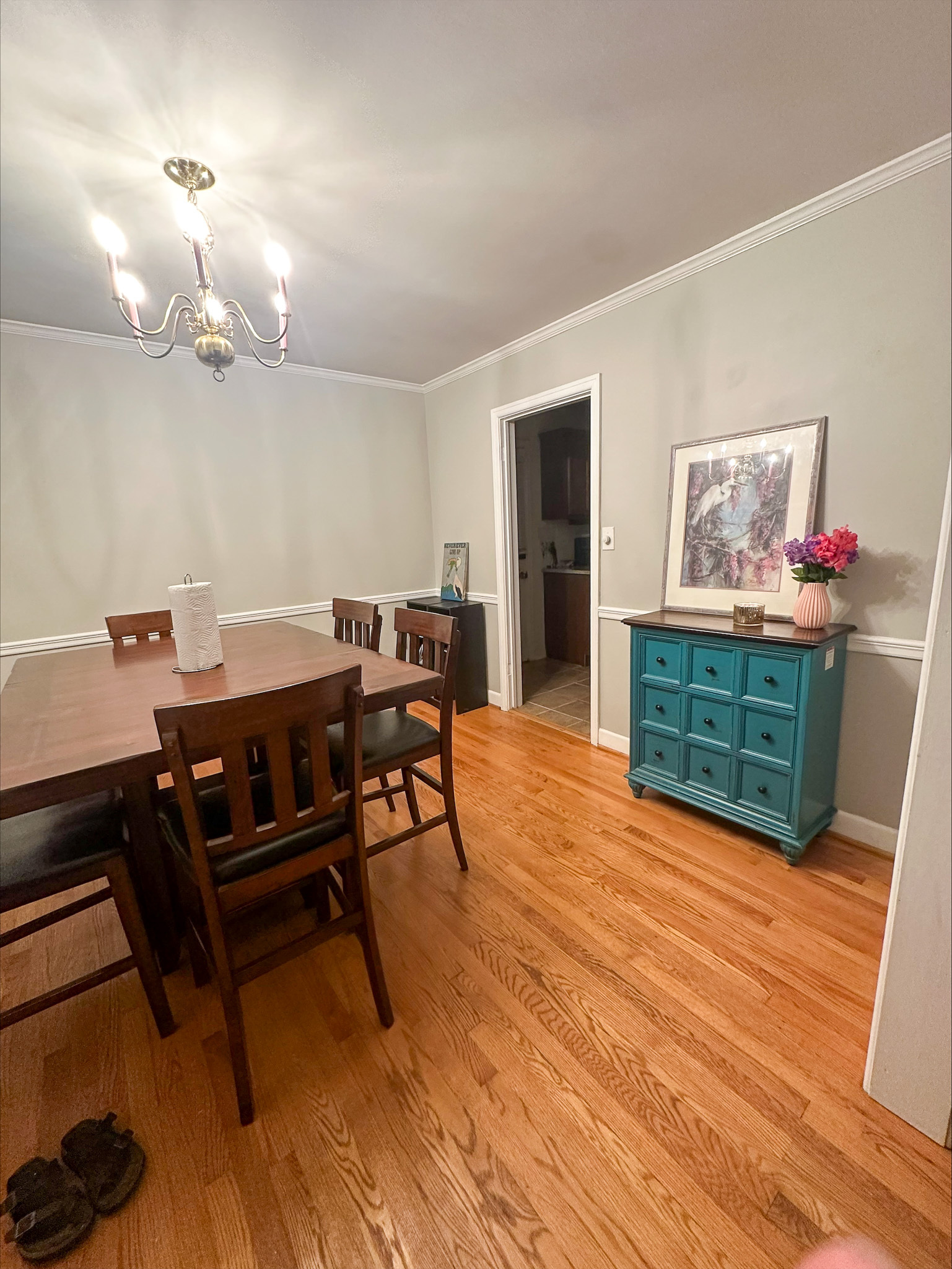 A dining room with a wooden table and chairs, hardwood floors, a chandelier, and a blue side cabinet with artwork above it inside the home of Group Home Provider Tina Langhorn in Roanoke, Virginia.