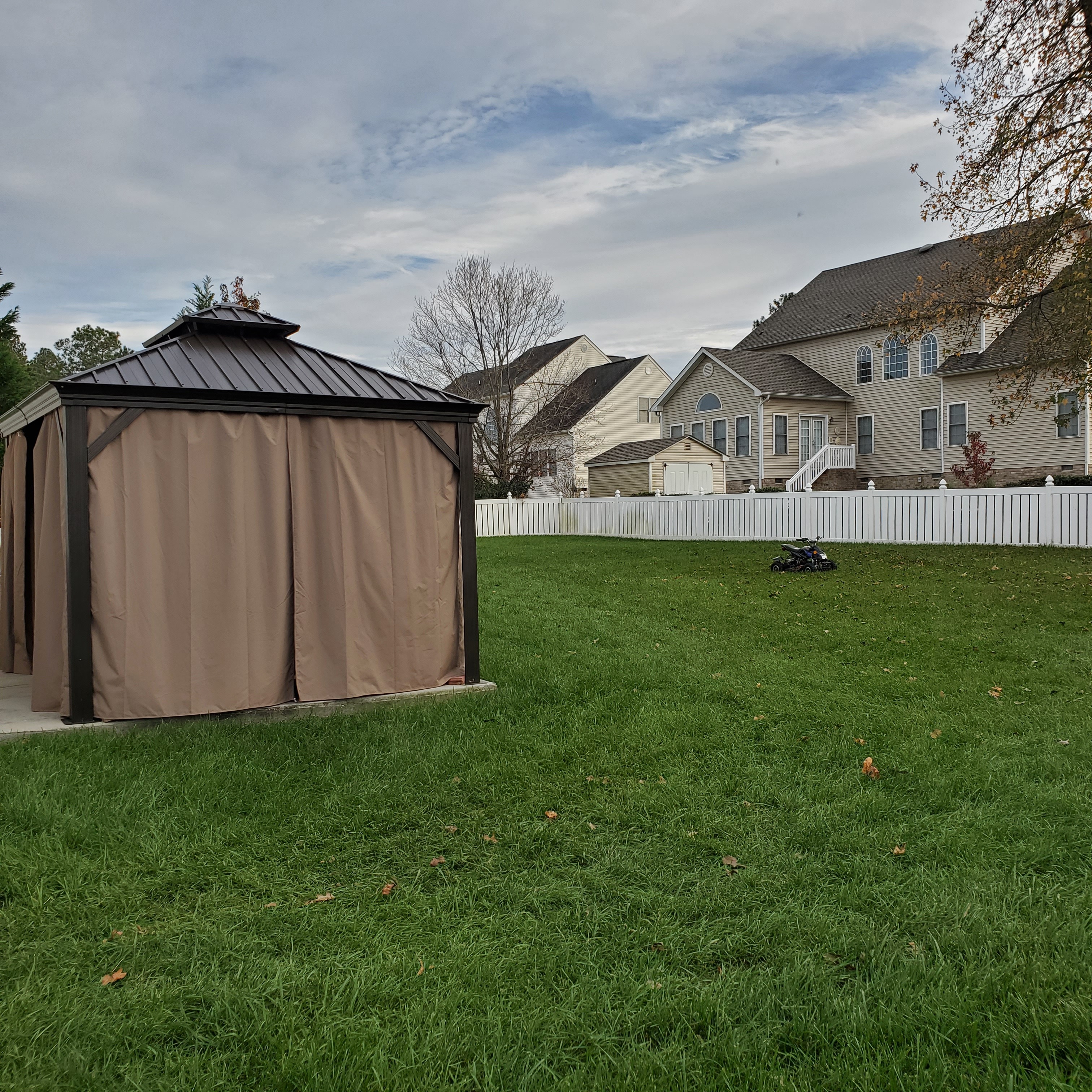 Backyard of home belonging to Group Home Providers Dayshawnna & Michael Lee with green grass, a covered gazebo with closed curtains on a concrete pad, and neighboring two‑story houses behind a white privacy fence.
