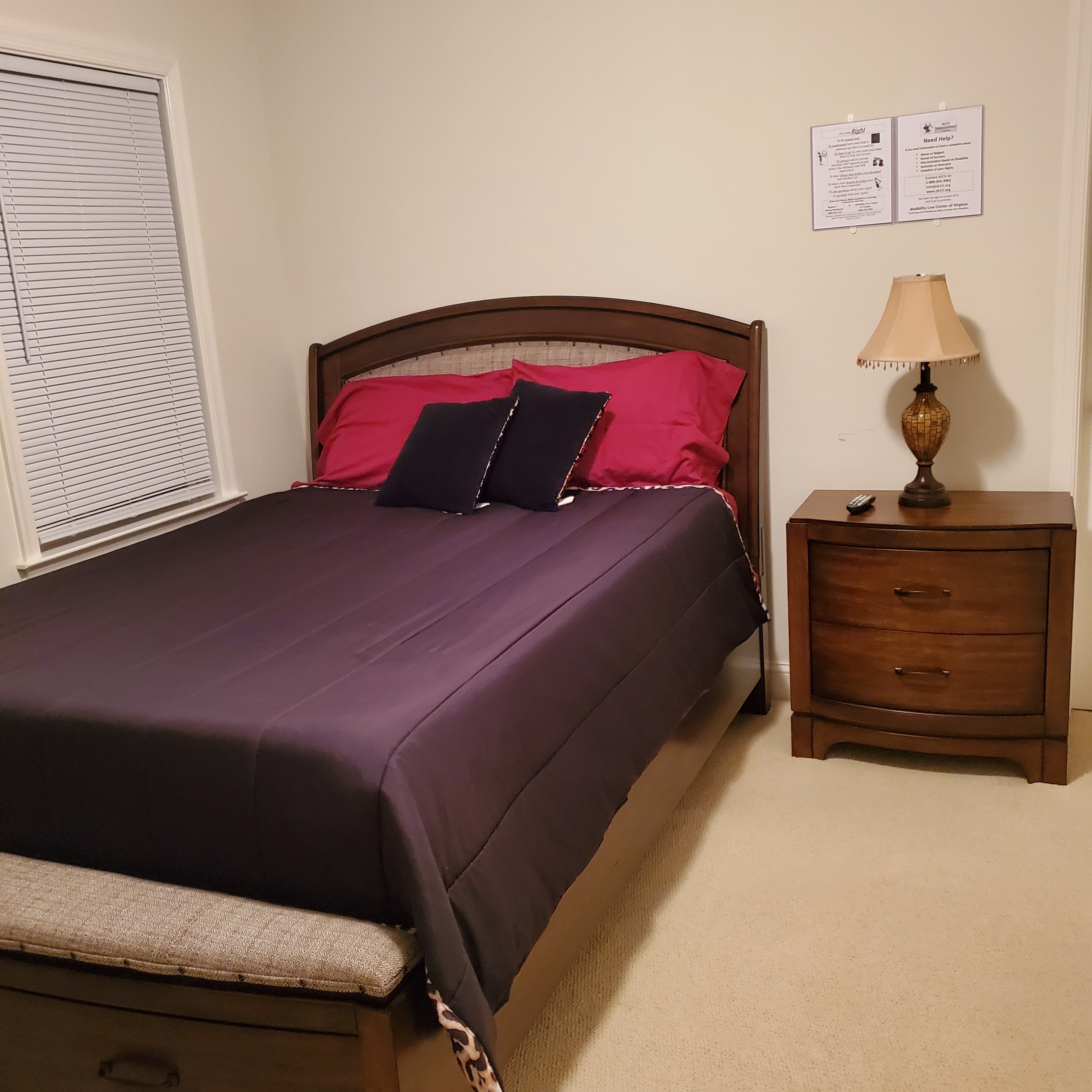 Bedroom with a full‑size bed dressed in a dark comforter and red pillows, a wooden headboard, a nightstand with a lamp, and closed blinds on the window inside the home of Group Home Providers Dayshawnna and Michael Lee in Chester, Virginia.