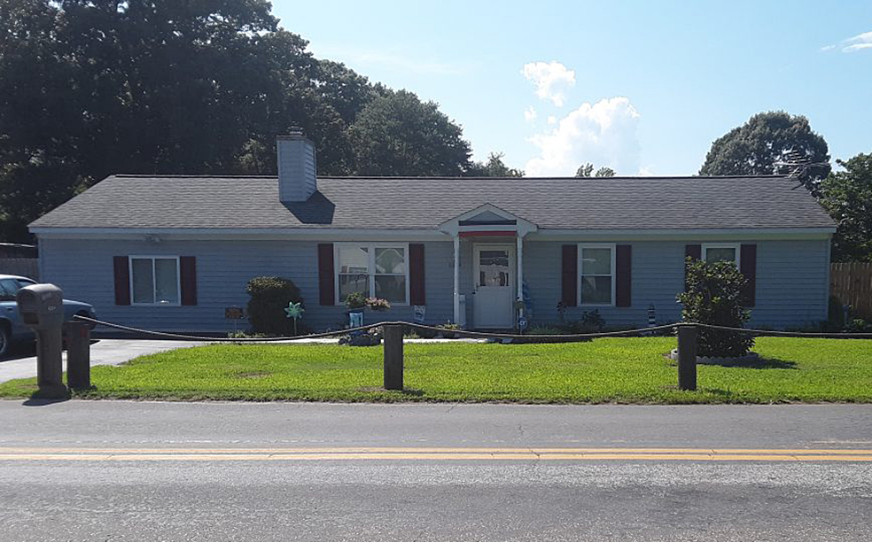 Single-story light-blue house with a gray roof, front porch, shutters, and a neatly mowed lawn along a roadside belonging to Group Home Provider Lori Cox in Hayes, Virginia.