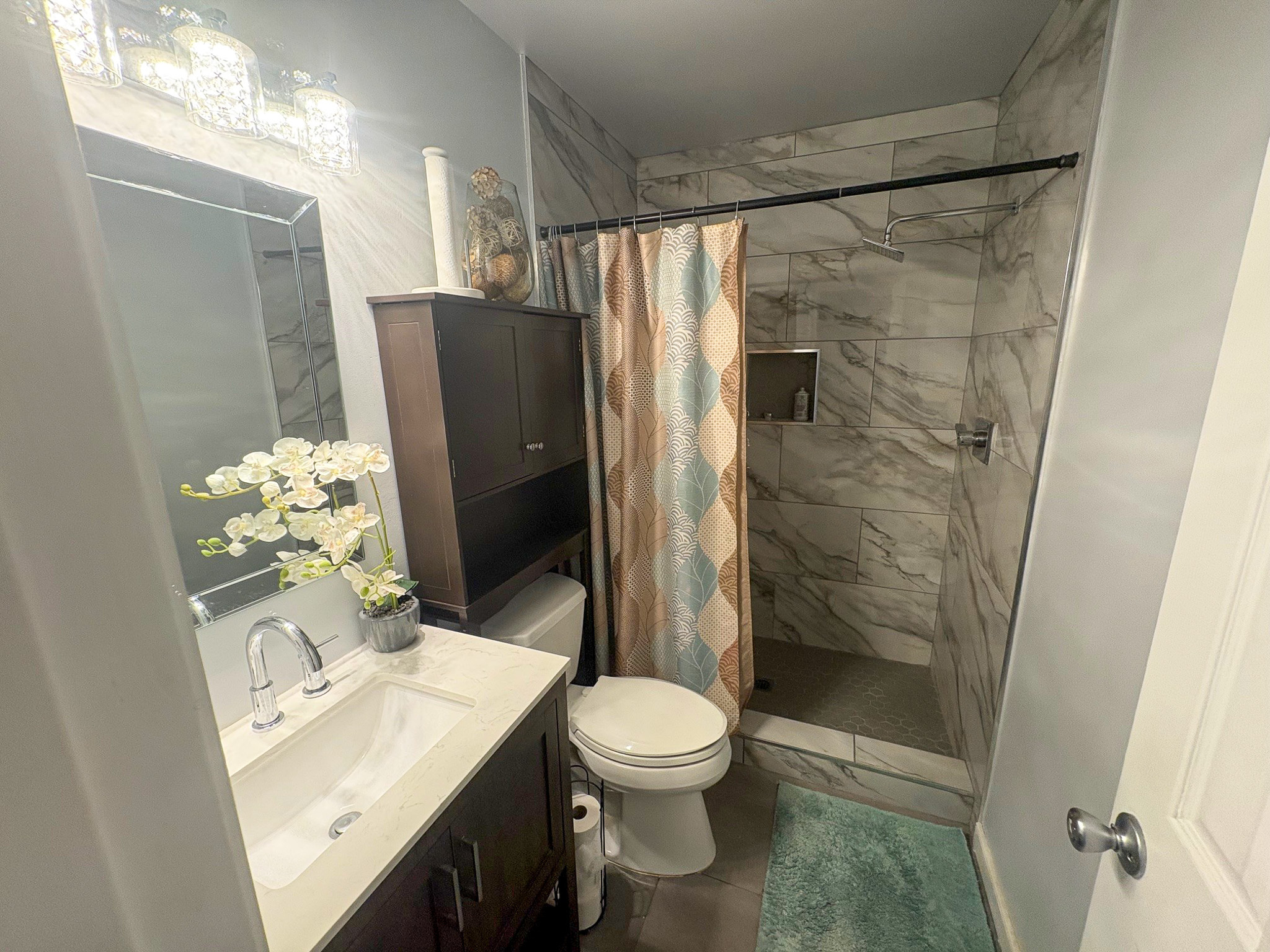 A bathroom features a vanity with a mirror, a toilet, and a tiled walk‑in shower with a patterned curtain and soft-colored accents inside the home of Group Home Provider Lelia Mabry in Midlothian, Virginia.