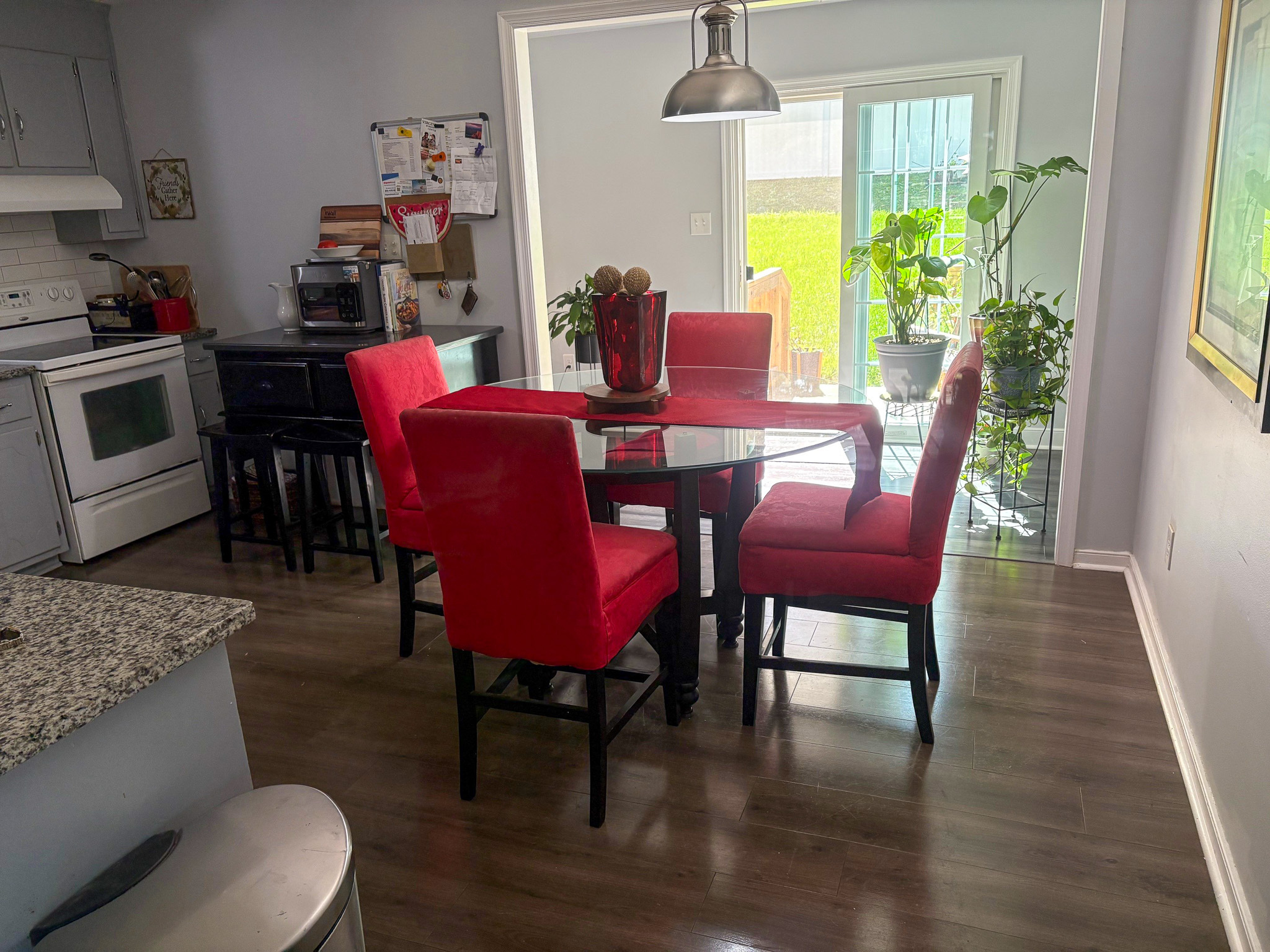 A dining area features a round glass table with bright red upholstered chairs, set beside the kitchen and a sunlit space with several potted plants inside the home of Group Home Provider Lelia Mabry in Midlothian, Virginia.