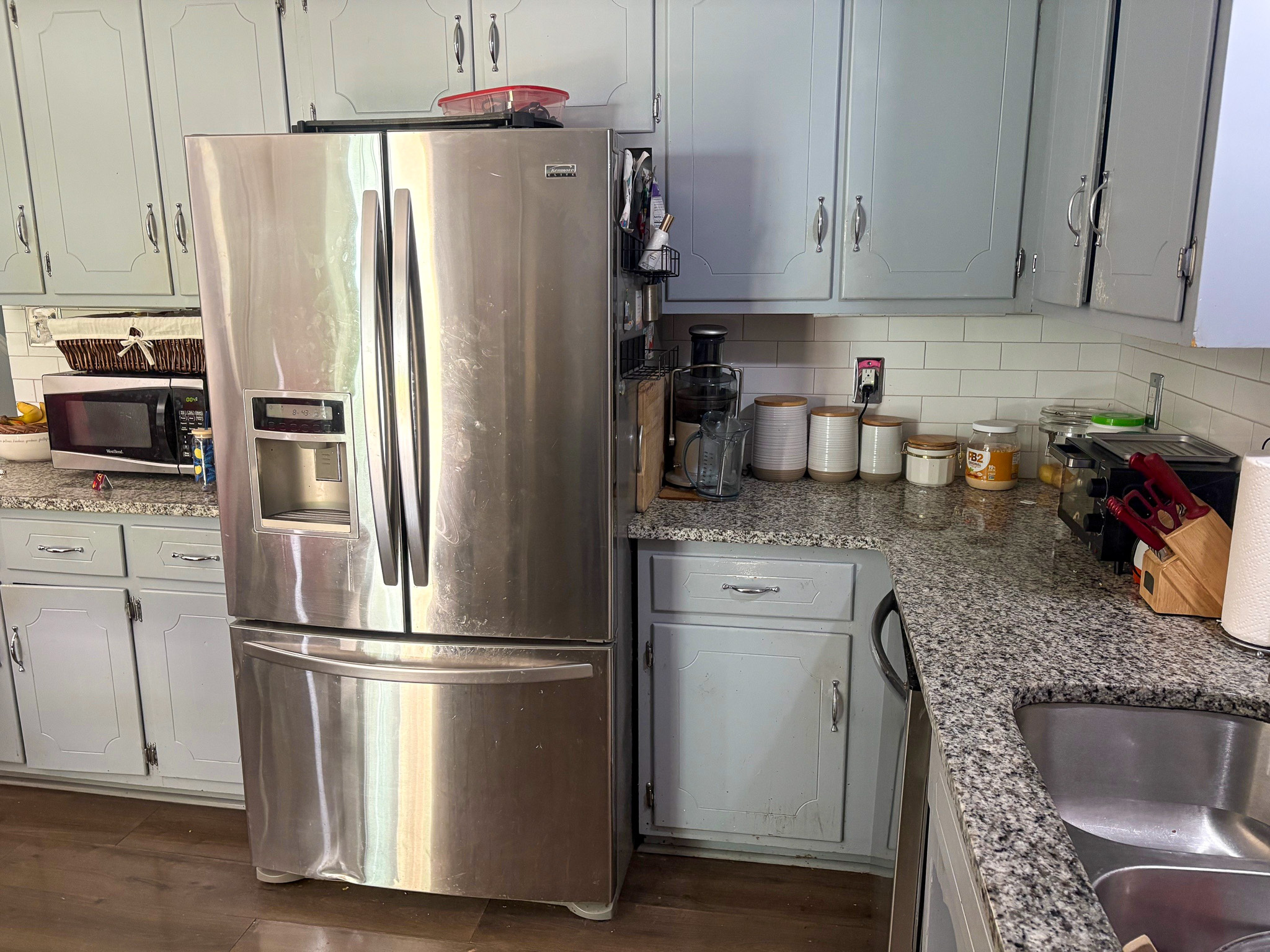 A kitchen counter with granite surfaces is lined with a stainless steel refrigerator, small appliances, and neatly arranged storage canisters beneath light‑colored cabinets inside the home of Group Home Provider Lelia Mabry in Midlothian, Virginia.