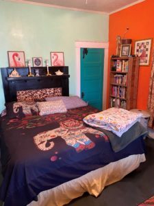Colorful bedroom with an elephant‑print bedspread, teal door, orange accent wall, and a tall bookshelf beside the bed inside the home of Sponsored Residential Provider Marcella Ajakaiye in Danville, Virginia.