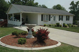 Single‑story white house with a covered carport, black shutters, concrete driveway, and a landscaped front yard with a birdbath and shrubs belonging to Group Home Provider Melissa Thompson-Daye in Danville, Virginia.
