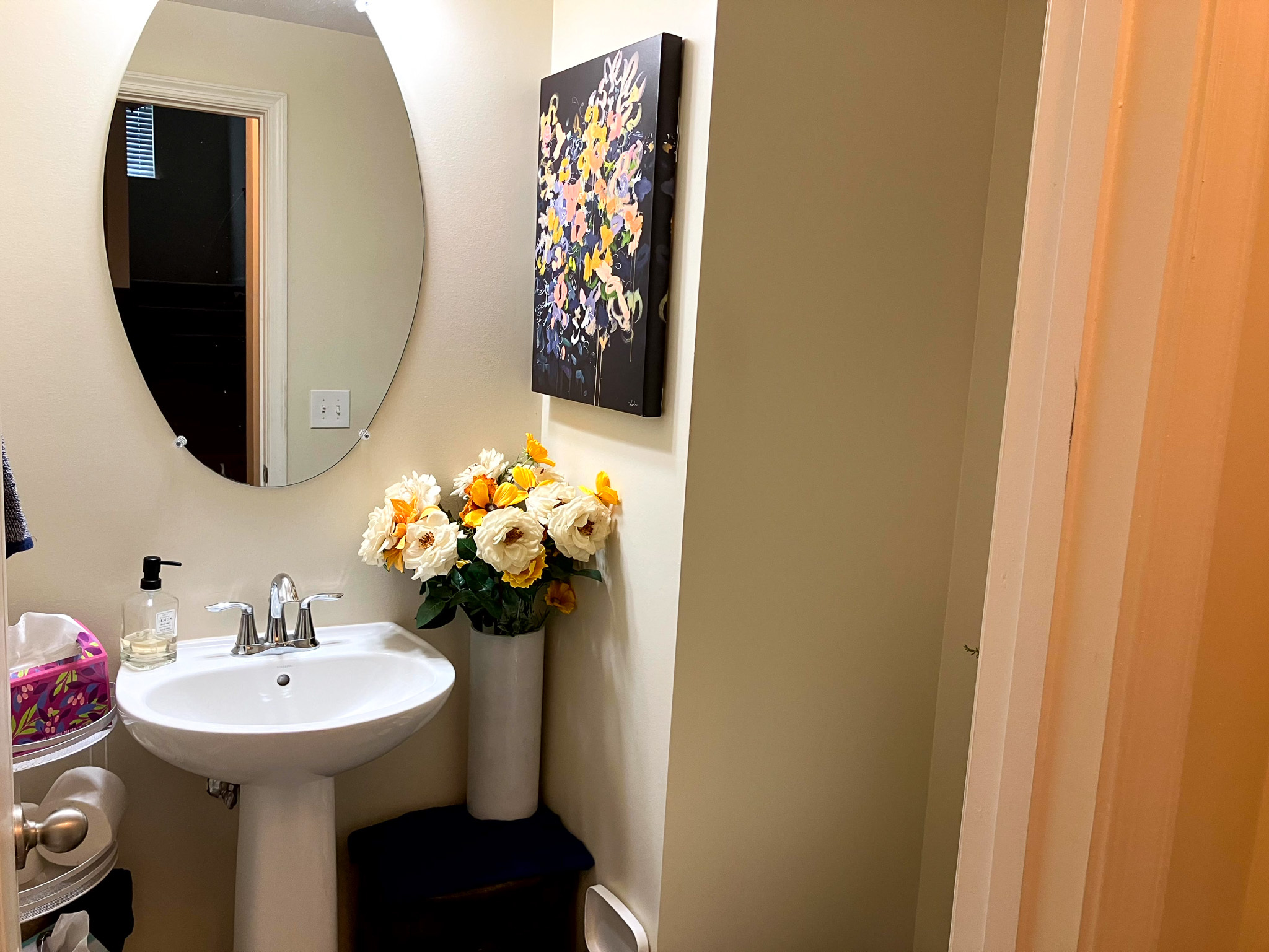A small bathroom with a pedestal sink, an oval mirror, floral wall art, and a tall vase of flowers beside the sink inside the home of Torwon and Deljuan Mitchell in Fredericksburg, Virginia.