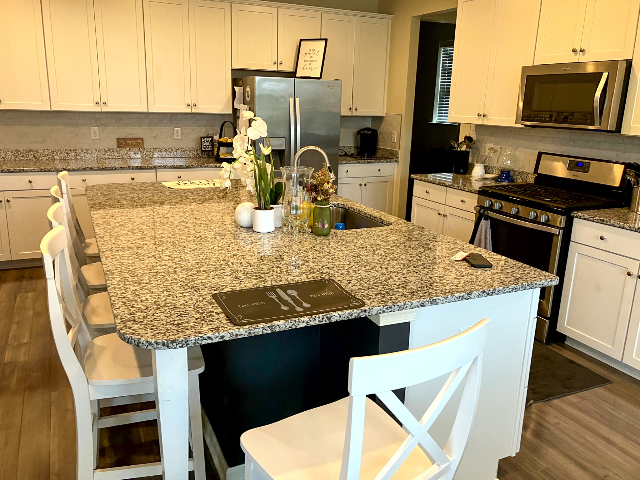 A modern kitchen with white cabinets, stainless steel appliances, and a large granite island with white barstools inside the home of Torwon and Deljuan Mitchell in Fredericksburg, Virginia.