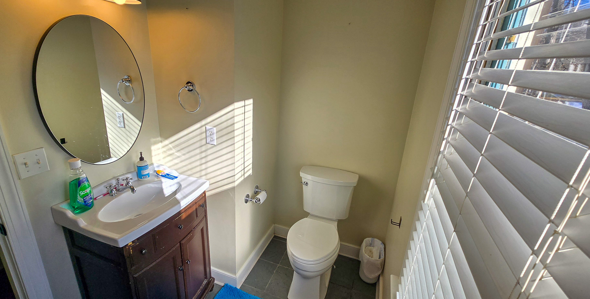 Small bathroom with a round mirror, dark vanity, toilet, and sunlight coming through window blinds inside the home of Group Home Provider Jared Nepal in Fredericksburg, Virginia.
