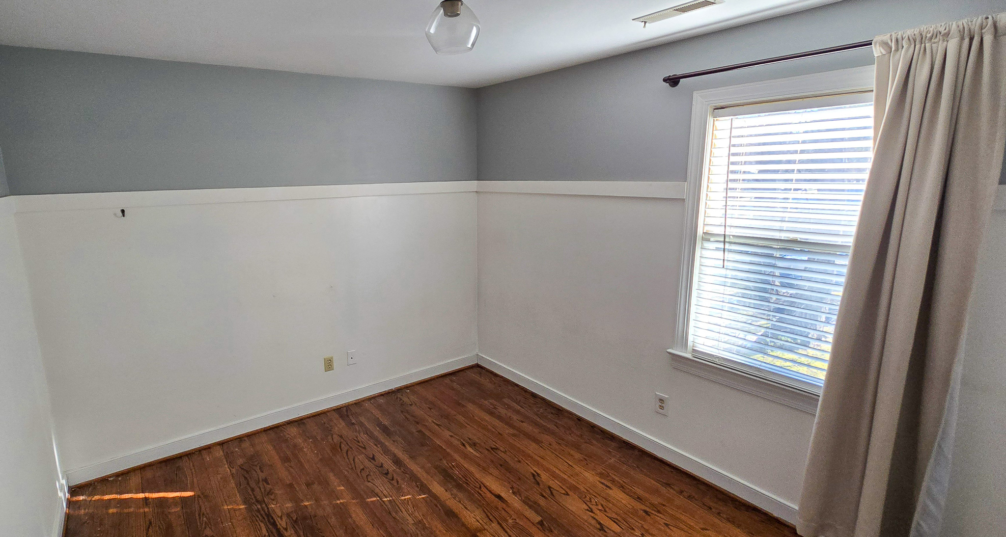 Empty room with gray and white walls, wood floors, a window with blinds, and a curtain on one side inside the home of Group Home Provider Jared Nepal in Fredericksburg, Virginia.
