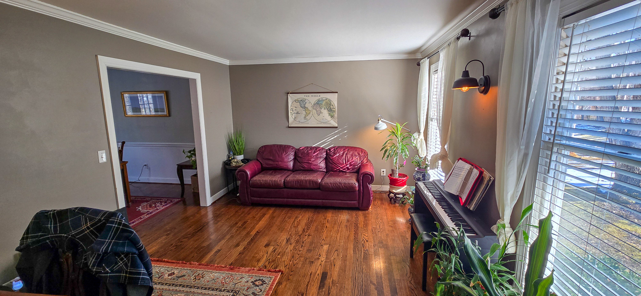Living room with a red leather sofa, wood floors, large window with blinds, several houseplants, and a piano along the right wall inside the home of Group Home Provider Jared Nepal in Fredericksburg, Virginia.